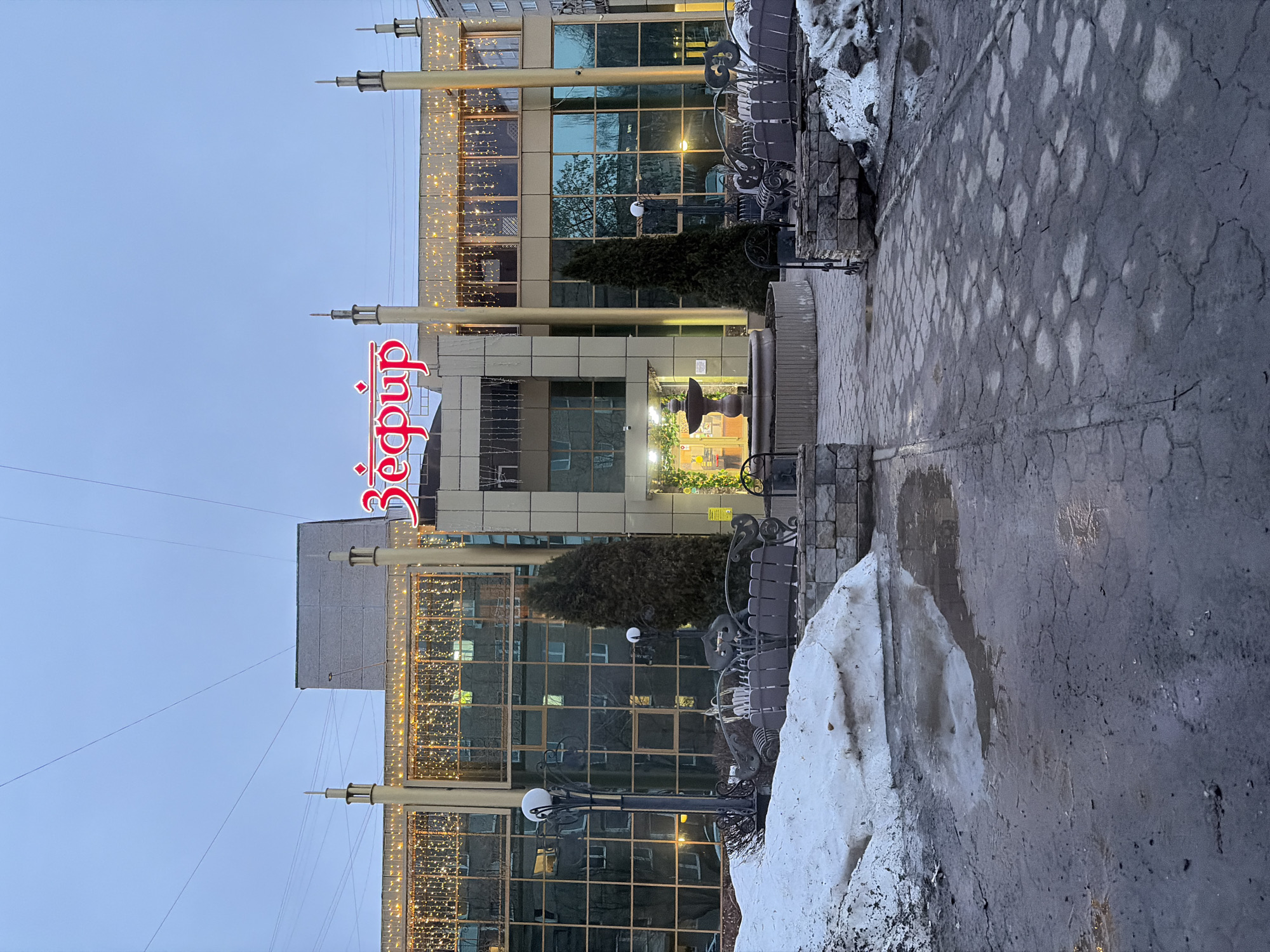 The image depicts a building exterior in Izhevsk, Russia. The building has a multistory facade with numerous windows and is adorned with strings of lights. A prominent red sign with Cyrillic lettering, reading "Вефир" vertically, is visible. At the building's base, there are benches and streetlights. Snow patches are present on the ground along the sidewalk. The sky is overcast. The focus is on the architectural details and urban environment.