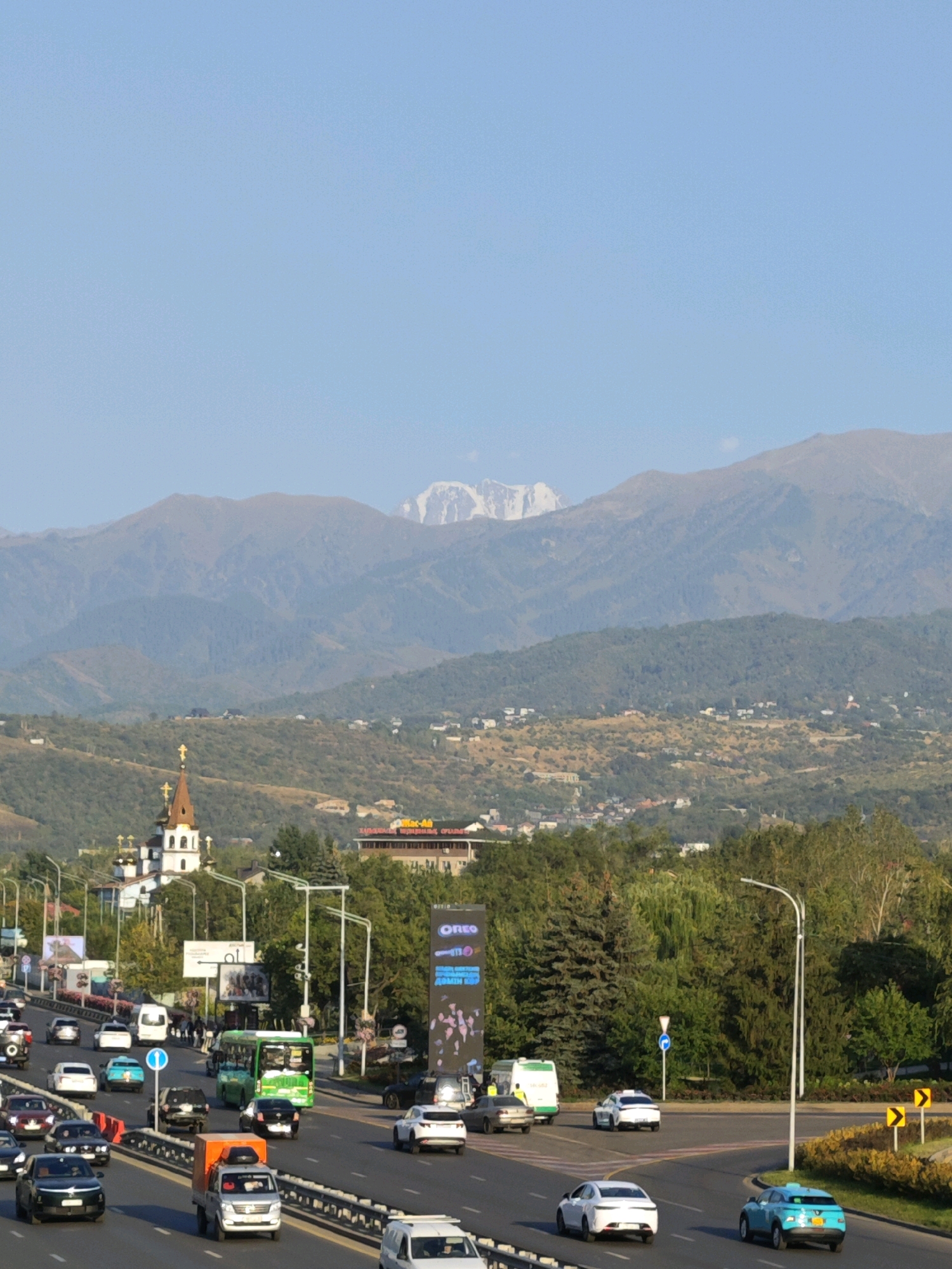 The image depicts a wide, multi-lane highway in Almaty, Kazakhstan, with a dense cityscape and mountainous backdrop. The foreground is dominated by a traffic jam on the highway, with numerous cars, trucks, and a green bus. A white van with an orange cargo container is in the center lane. Pedestrians are visible on a sidewalk to the left.
In the middle ground, several buildings are present, including a white church with a prominent spire topped with a cross. Adjacent to the church, a large billboard with the word "Oreo" is visible. Dense greenery, including trees and shrubs, fills the space between the highway and the distant hills.
The background features rolling, treed hills that transition into rugged, snow-capped mountains under a clear blue sky. The overall impression is of a bustling urban environment situated at the foothills of a majestic mountain range.