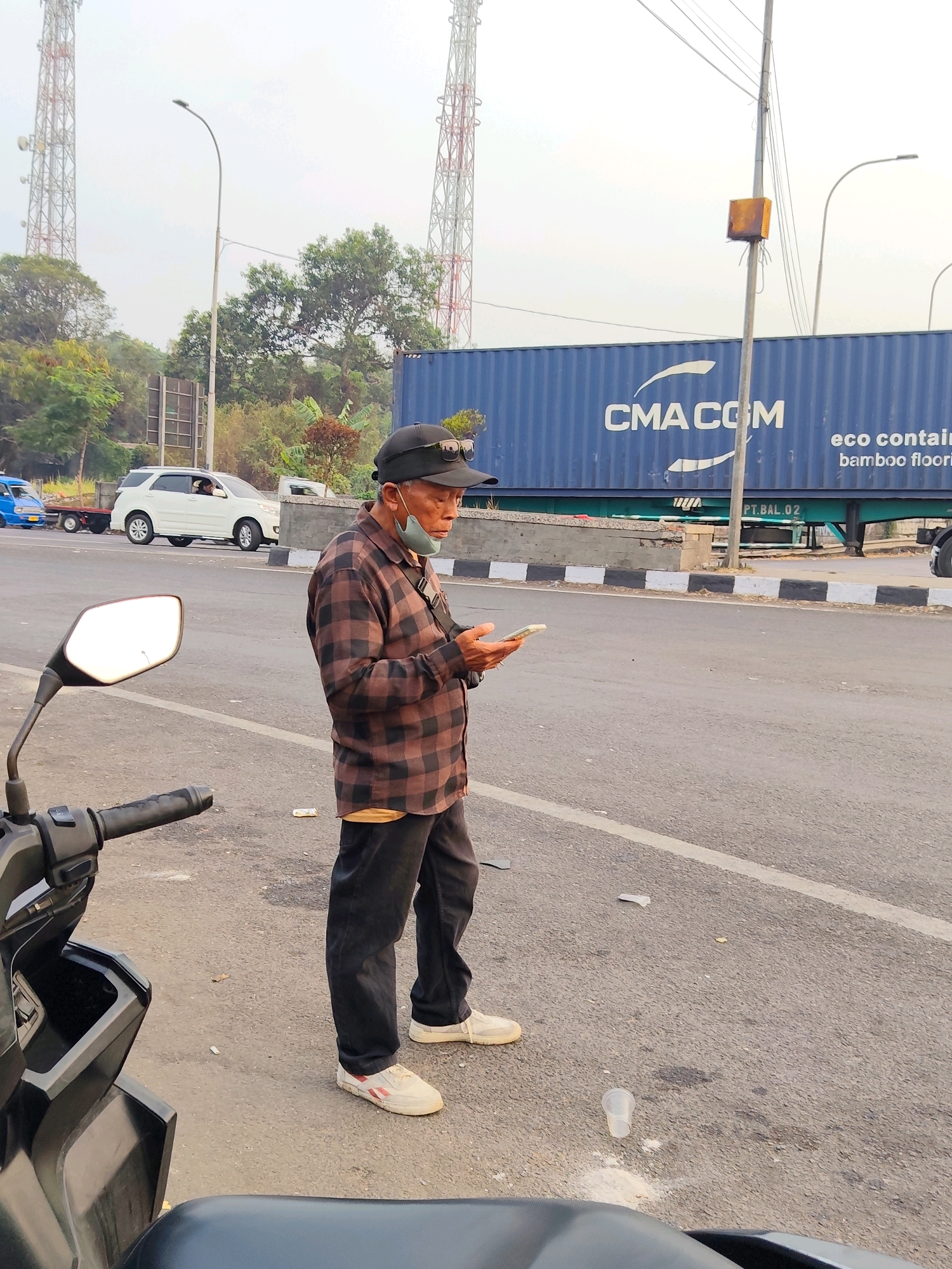 An elderly East Asian man wearing a plaid shirt, black pants, a black baseball cap, sunglasses, and a light blue surgical mask pulled down below his nose stands on the side of a road in Cikampek, Indonesia. He is looking at a mobile phone in his hands. In the foreground, the side mirror and handlebar of a black motorcycle are visible. Behind the man, a white SUV is parked on the road, and a blue truck with a trailer is driving away. A large blue shipping container with "CMA CGM" branding is visible in the background, along with telecommunication towers. The ground is paved with asphalt and appears to be a roadside. The sky is overcast and bright. There are some pieces of litter on the road.