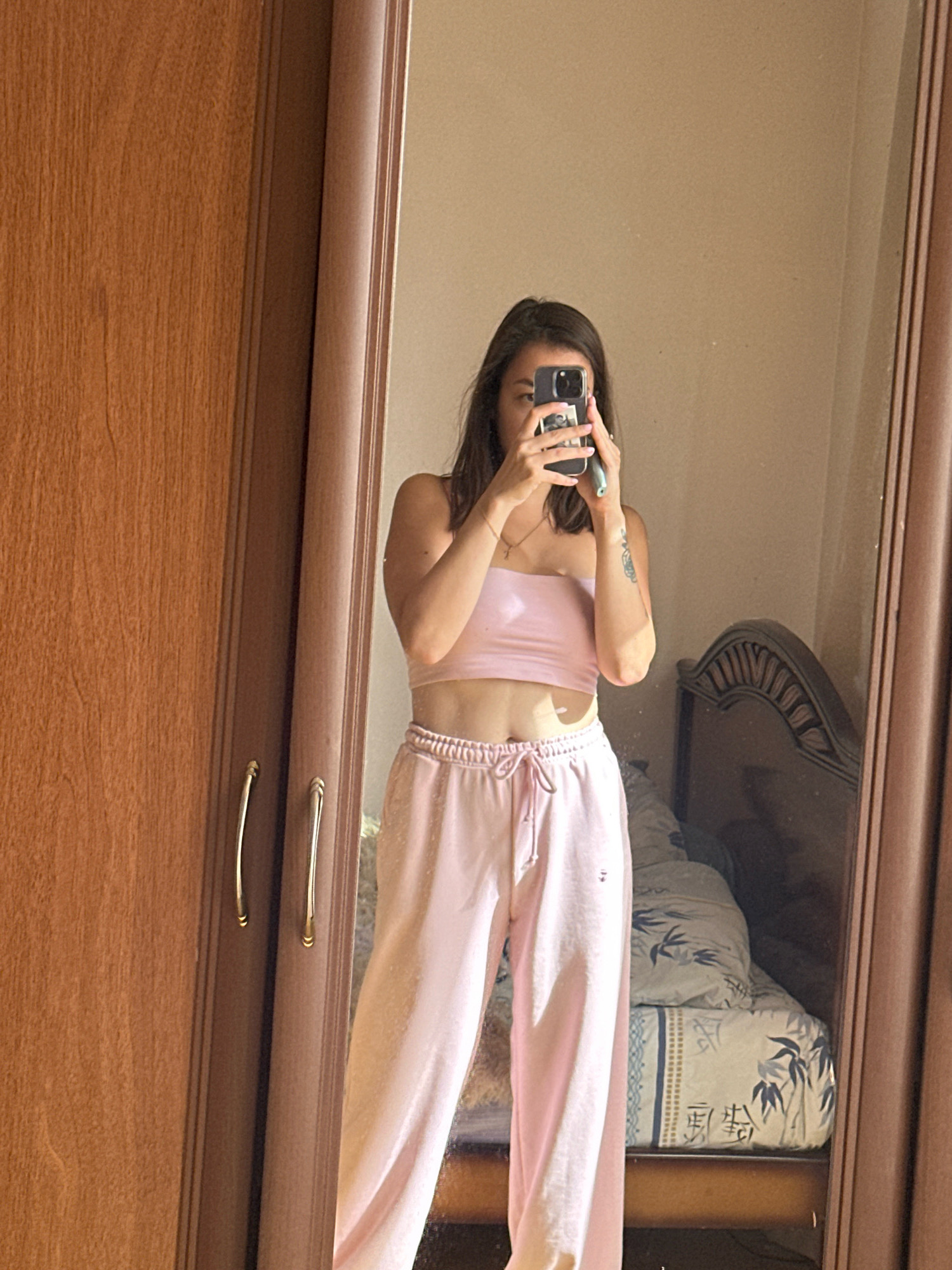 In Gyumri, Armenia, a young woman stands in front of a full-length mirror, taking a selfie with her smartphone. She is wearing a light pink strapless top and matching pink sweatpants. Her dark hair is down, and she has a tattoo on her right arm. The mirror reflects part of a bedroom, including a bed with a patterned comforter and a dark wooden headboard. To the left of the mirror is a tall, wooden wardrobe with two doors and brass handles. The lighting suggests it is daytime, with sunlight casting shadows.