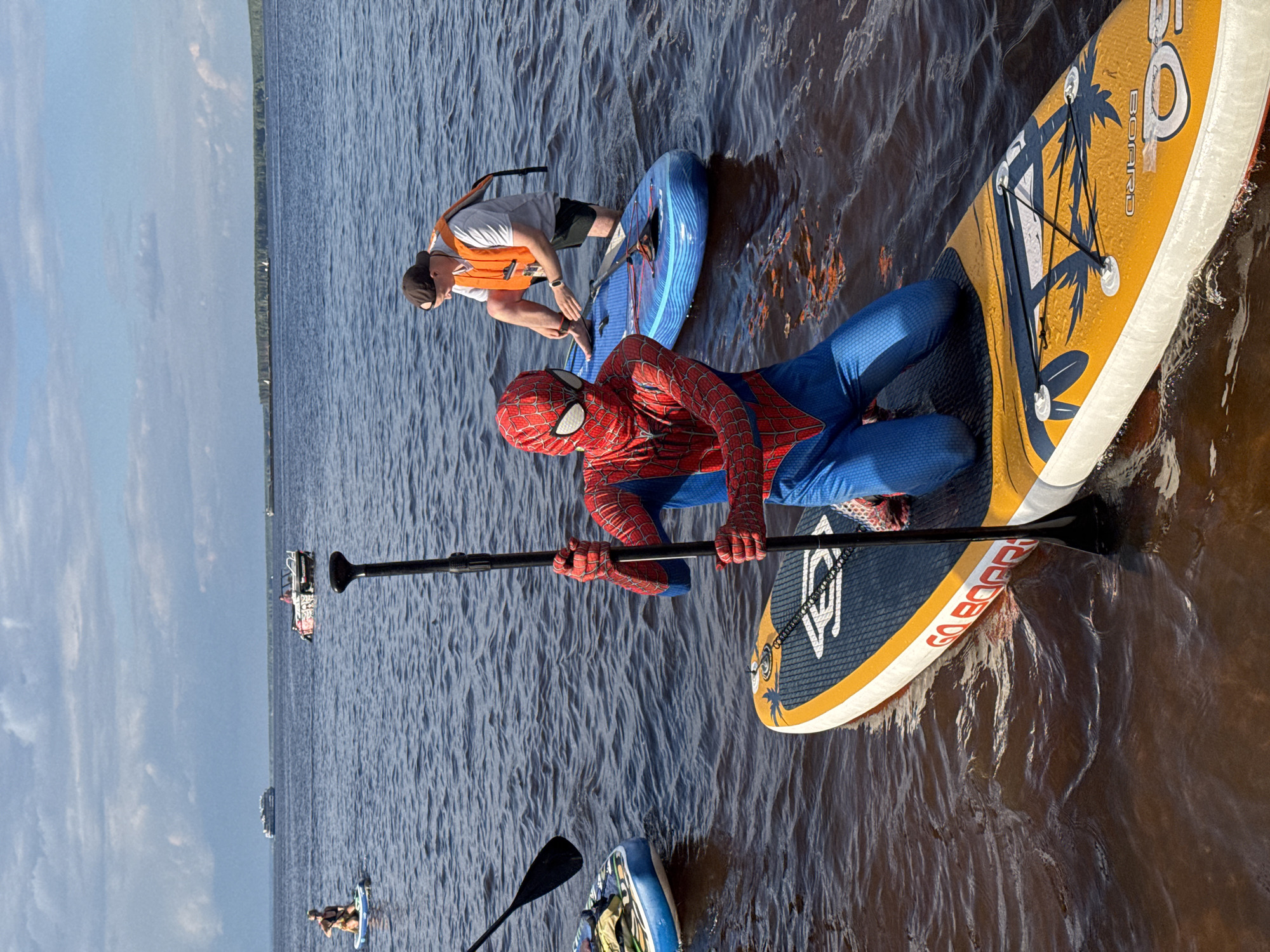 A person in a Spiderman costume kneels on a yellow and blue stand-up paddleboard in a body of water, holding a paddle. Another person wearing an orange life vest and a baseball cap is standing in the water next to a blue paddleboard, tying their shoe. In the background, there are other paddleboards and boats on the water, with a forested shoreline and a cloudy sky. This scene is taking place on the Kama River in Chaikovsky, Russia.