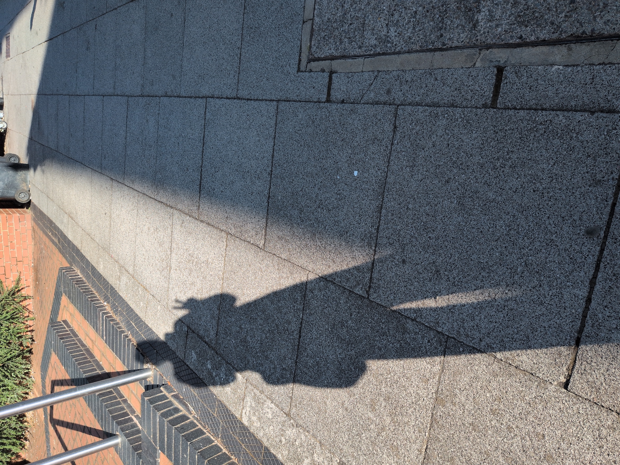 The image shows a shadow of a person casting on a paved sidewalk. The person is standing, and their shadow is elongated, indicating a low sun angle. The shadow shows a figure with their left arm raised, fingers extended, possibly making a peace sign or waving. The environment includes a brick wall and some greenery to the left, as well as a set of metal railings and steps. The sidewalk is made of large, grey, speckled paving stones. Part of the sidewalk is in sunlight, and the other part is in shadow. There is a metal grate in the lower right corner of the image. There are no people visible in the image, only the shadow of one person. The location is the city of Dawley, United Kingdom.