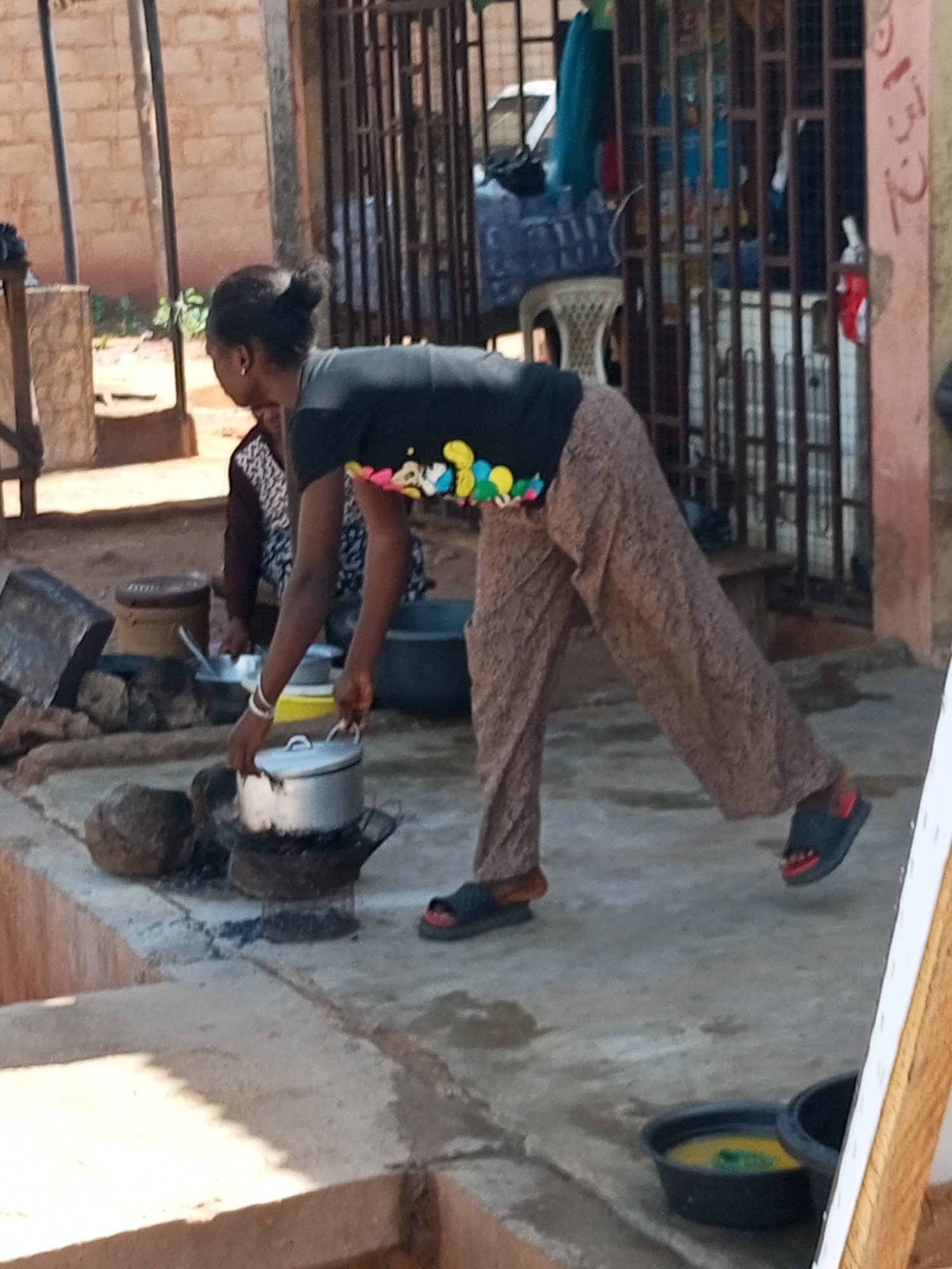 The image captures an outdoor scene in Tempure, Nigeria, with a woman actively engaged in cooking. She is dressed in a black t-shirt with a colorful, patterned design around the collar and loose, patterned brown trousers. Her right hand is shown reaching for or adjusting a silver pot that sits on a makeshift stove constructed from dark stones. To the left of the stove, another woman, partially obscured, is seated and appears to be involved in a similar activity, with a patterned garment visible. The environment suggests a humble dwelling or marketplace, characterized by a dirt or concrete ground surface, brick walls in the background, and a metal grate structure. Several cooking vessels, including pots and what appears to be a metal basin, are scattered around the immediate cooking area. The lighting indicates it is daytime, casting shadows on the ground.