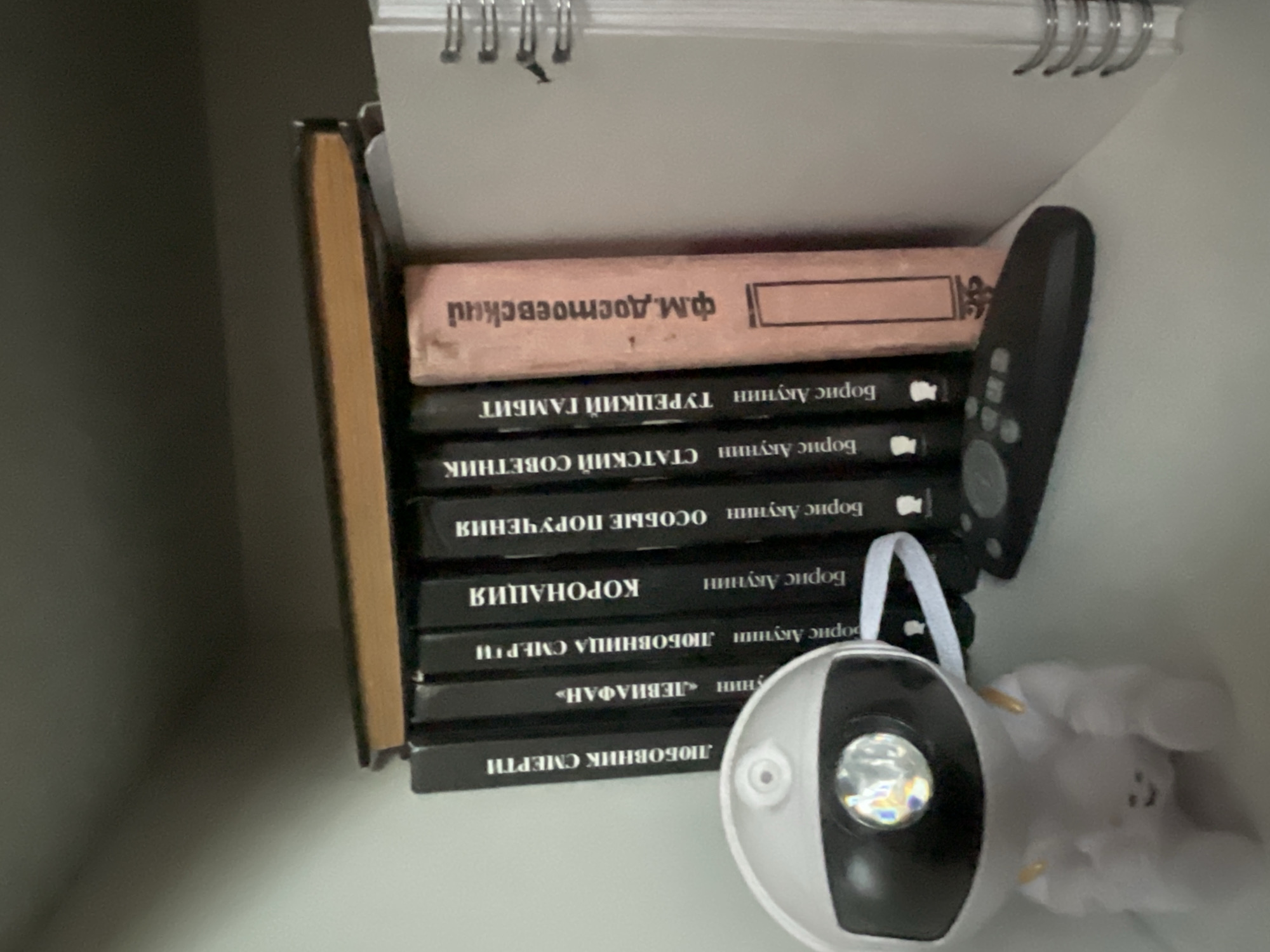 The image displays a shelf in a room, likely a home or office, with several books and a small white robot-like figurine. The books are arranged vertically, with their spines facing outwards. The titles on the spines are partially visible, appearing to be in Russian. One book spine reads "ЛЮБОВНИК СМЕРТИ" (Lover of Death), another "ЛЕВИНАФАН" (Leviafan), and another "КОРОНАЦИЯ" (Coronation). To the right of the books is a white, spiral-bound notebook. A remote control rests on the shelf in front of the books, and the white figurine is positioned to the left, facing forward with a light on its head. The background is a plain, light-colored wall. The overall impression is of a neatly organized shelf. The location is identified as Мухавецкі сельскі Савет, Belarus.