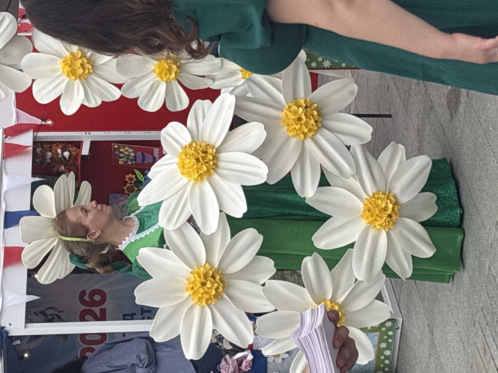 The image depicts an outdoor scene, likely a festival or market, with a woman in a green dress standing amidst large, artificial white and yellow flowers. She has her hair tied back with a green ribbon and is looking to her right. To her left, a person in a blue striped shirt is partially visible. In the background, a red and white striped structure is visible, along with other decorative elements, including a banner with the text "2026". The ground appears to be paved with grey interlocking stones. The scene is colorful and festive. The context suggests it might be a public event in Astrakhan, Russia, given the presence of the year "2026" which could be related to an upcoming event or anniversary.
