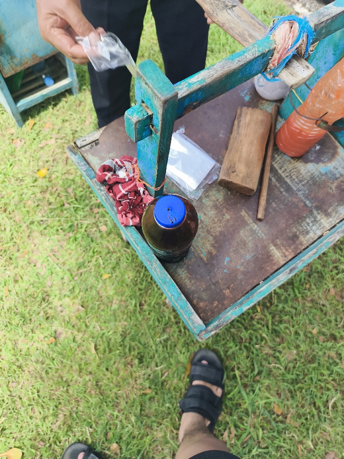 A person is standing next to a wooden cart. The cart is painted blue and green and has a variety of items on it including a glass bottle with a blue lid, a red and white cloth, a wooden block, a plastic bag, a clear plastic bag, and a blue rope. The cart is sitting on green grass. The location is in Majalengka, Indonesia, at the coordinates provided (latitude: -6.846, longitude: 108.204). The person is wearing dark shorts and sandals. The person's hand is reaching into a plastic bag, and the person's left foot is resting on the ground.
