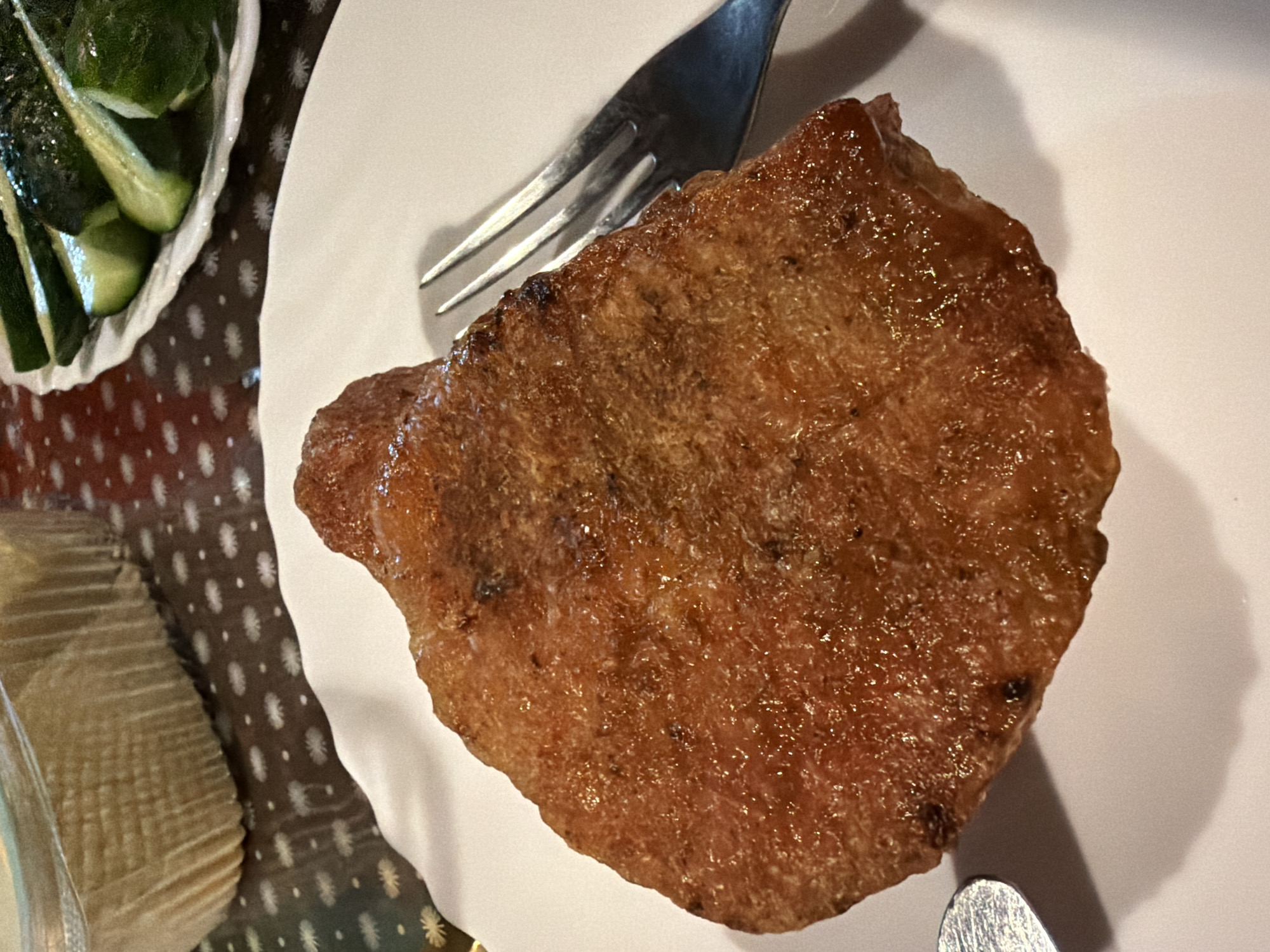 A close-up, top-down view captures a golden-brown, fried food item, possibly a cutlet or piece of meat, resting on a white plate. A fork is positioned to the right of the food, its tines pointing towards the center of the plate. The background features a patterned tablecloth in shades of brown and white, with a portion of a white, ribbed container visible in the upper left corner. In the upper right, a plate holds sliced cucumbers. The lighting suggests an indoor setting, likely a restaurant or home. No people are present, and no specific location in Sochi, Russia, is identifiable from the image.
