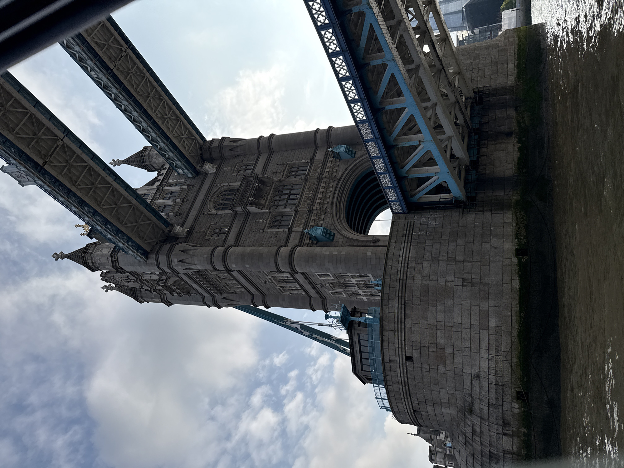 The image depicts the Tower Bridge in London, United Kingdom. The bridge's towers, made of gray stone, feature arched openings and decorative elements. The bridge's upper structure is made of steel and painted blue and gray. The River Thames flows alongside the bridge's foundation. The sky is visible through the bridge structure, with scattered clouds.