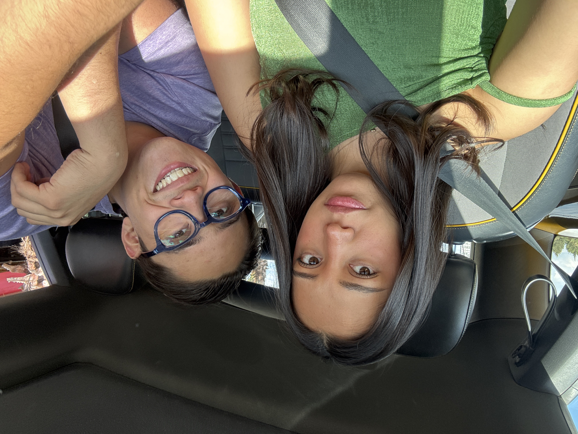 Two people, a male and a female, are inside a vehicle. The male is wearing glasses and a purple shirt, and the female is wearing a green top and a seatbelt. The location cannot be determined as San Bernardino Tlaxcalancingo, Mexico based on the visual data provided. The environment appears to be inside a car.