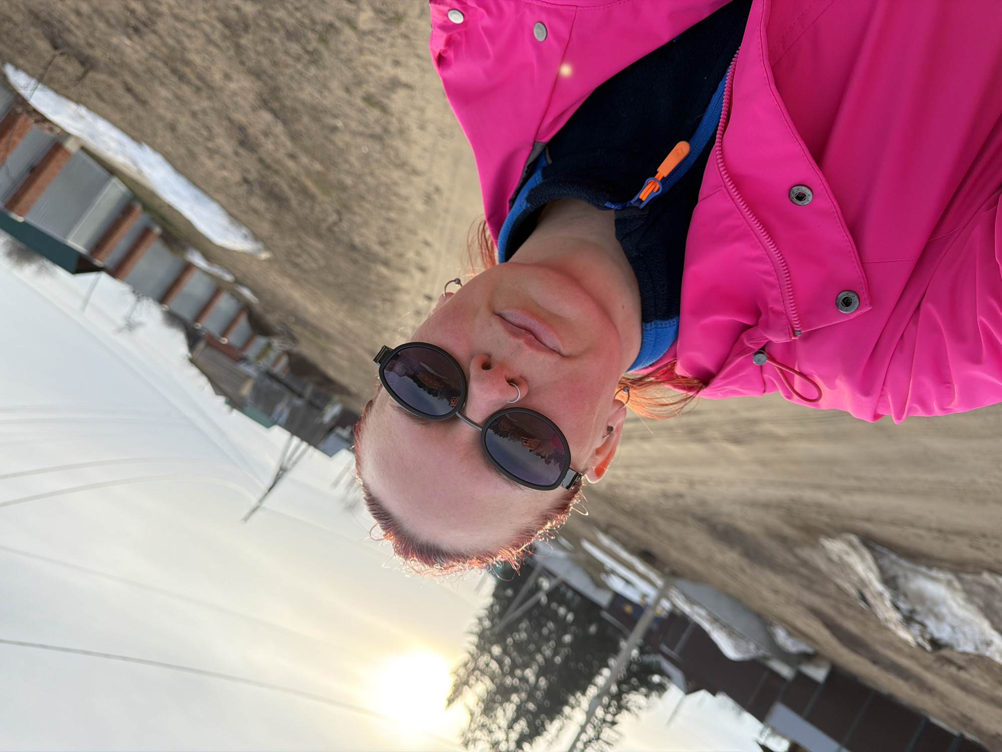 A woman wearing a bright pink jacket and dark sunglasses is in an outdoor environment. She has a nose ring and earrings. The background shows houses with snow patches and what appears to be a field. The sun is setting or rising, casting a bright light. The image appears to be taken near the city of Chaikovsky, Russia, suggested by the snow-covered landscape.