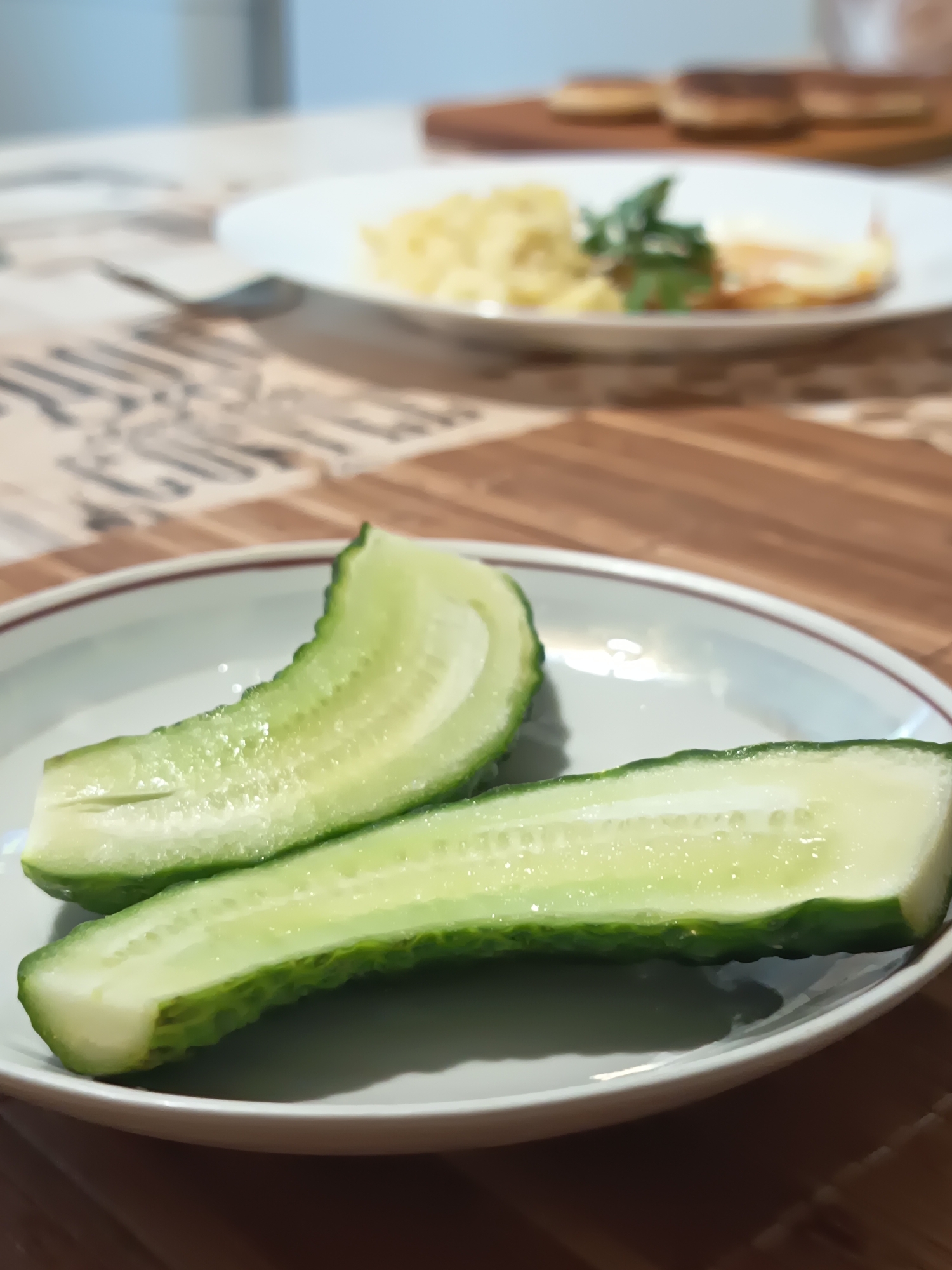 Two halves of a cucumber are presented on a white plate with a dark rim. The cucumber pieces are cut lengthwise, revealing their seeds and watery interior. The plate sits on a wooden placemat with a textured, patterned surface. In the background, a blurred plate of food, possibly pasta and a fried egg, is visible on a table. The overall scene appears to be a domestic setting, likely a meal being prepared or served. There is no identifiable location in Izmail, Ukraine.
