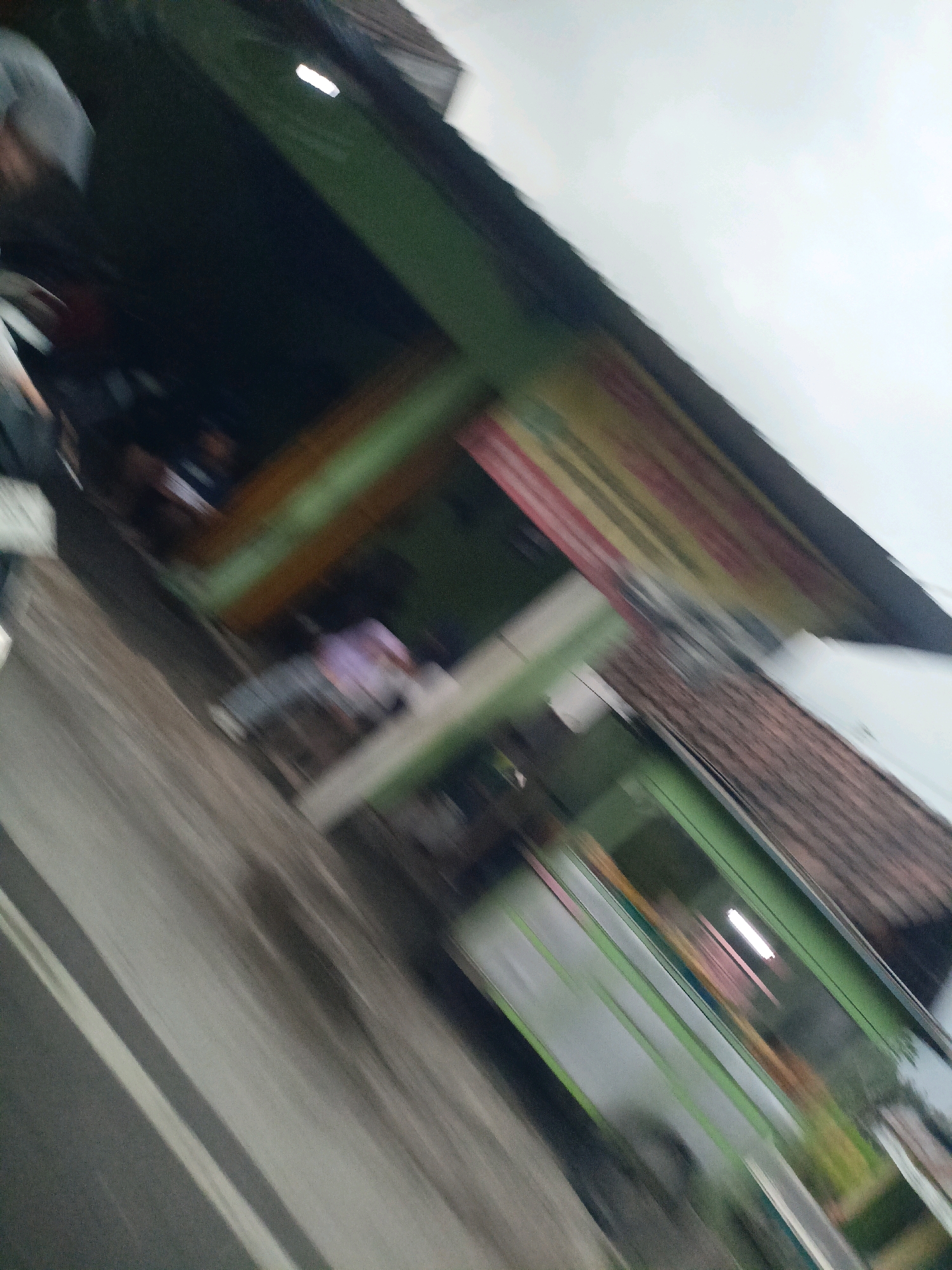 The image is a motion-blurred photograph taken from a vehicle traveling along a street in Payaman, Indonesia. The scene depicts a row of buildings with green and brown facades, and a section of a tiled roof is visible. A person in a light-colored shirt and dark pants is walking on the sidewalk in front of one of the buildings. The road in the foreground is wet and appears to have white lane markings. The sky is overcast.
