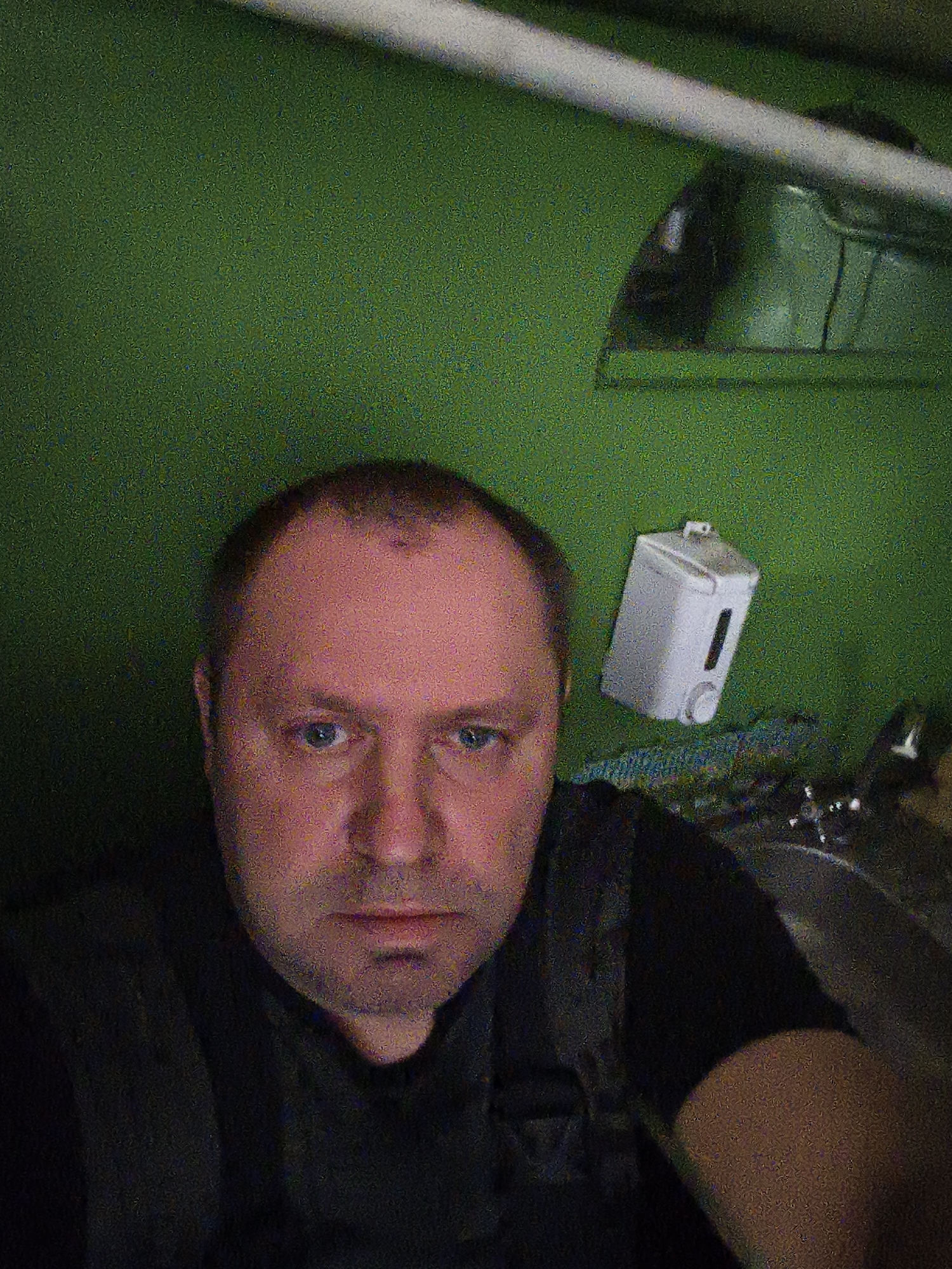 A medium shot shows a man in the foreground, facing the camera. He has fair skin, blue eyes, and a shaved head. He is wearing a dark shirt and what appears to be dark overalls. The man is positioned in front of a vibrant green wall, which takes up most of the background. To the right of the man, mounted on the green wall, is a white dispenser, possibly for soap or sanitizer, with a black vertical indicator. A silver pipe runs horizontally across the top of the frame, above the green wall. The lighting appears to be artificial, creating a somewhat grainy texture throughout the image. The scene does not contain any recognizable landmarks or clues that would indicate a specific location within Daugavpils, Latvia.