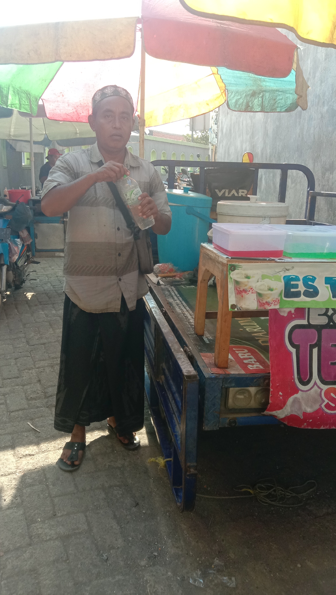 A man wearing a prayer cap, a striped short-sleeved shirt, and a black sarong is standing in front of a cart with a blue frame and a blue cooler. He is holding a plastic bottle of water, with the cap loosened. The cart is adorned with signs, one featuring images of iced drinks and the text "ES TEH". The scene is outdoors, with bright sunlight casting shadows on the paved ground. The background shows a building with a gray wall and a partially visible street with parked motorcycles. The environment appears to be a marketplace or roadside stall.