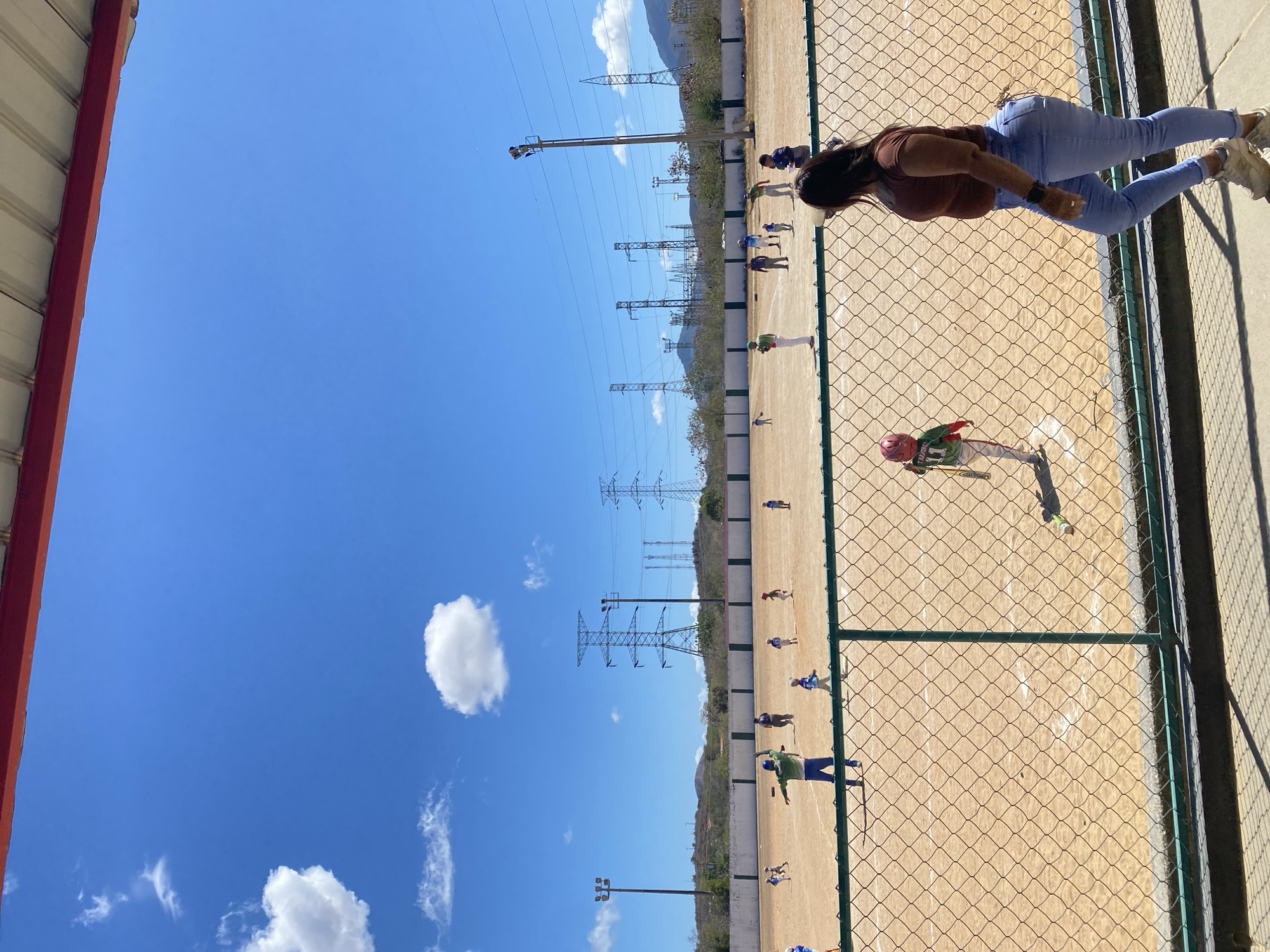The image captures a baseball field scene, likely in Juan Griego, Venezuela, given the mention of the city. A woman wearing blue jeans and a brown shirt is seen standing near a green chain-link fence, possibly observing a baseball practice or game. A young baseball player in a red helmet and green uniform is in action on the field. Other baseball players in various colored uniforms can be seen dispersed on the field. Overhead power lines are visible in the background. The sky is blue with white clouds.