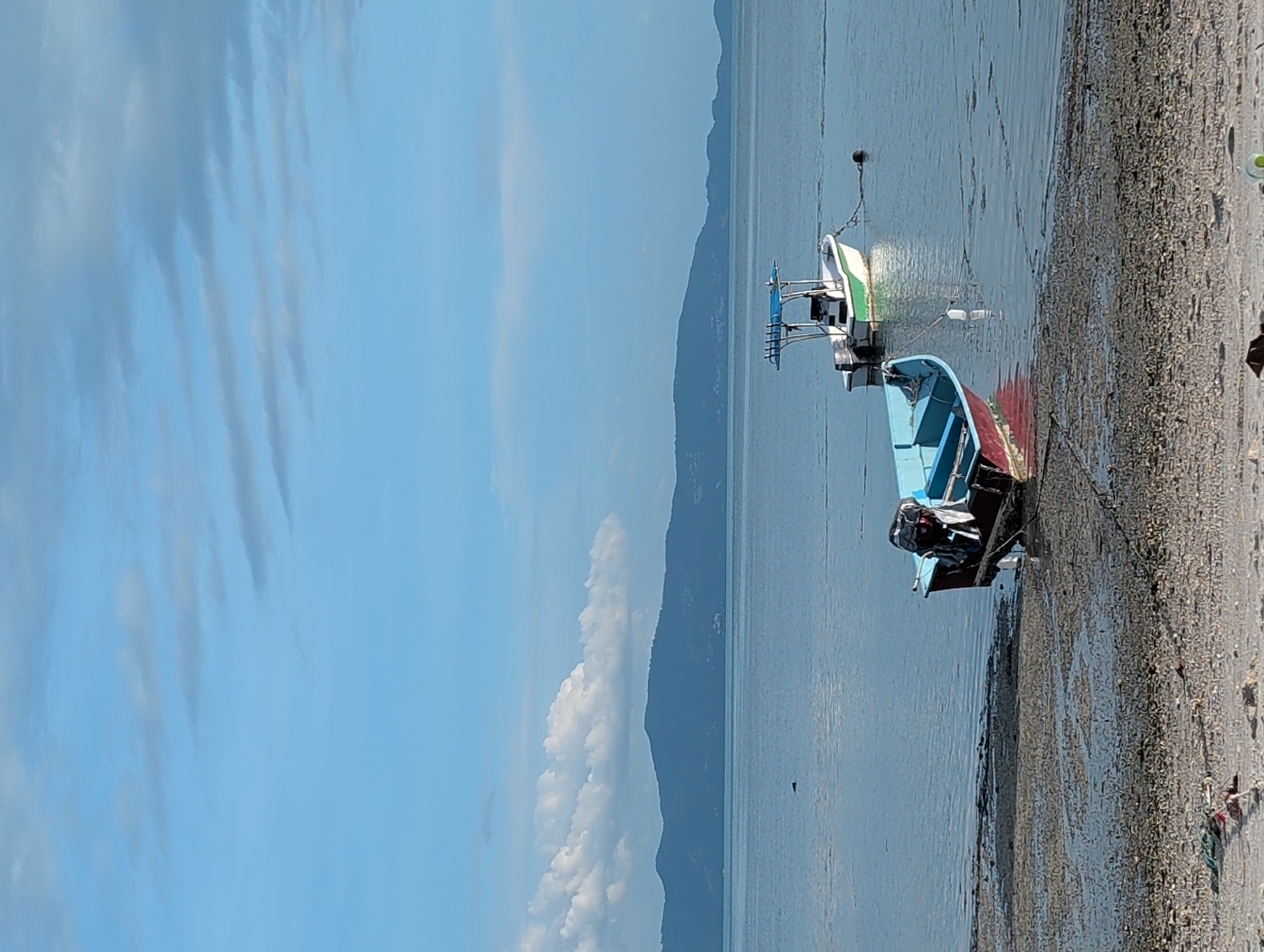 The image depicts a coastal scene in ตำบลเกาะพะงัน, Thailand. Two boats are present: one is beached on the wet sand with a motor, and the other is floating further out in the water. A mountain range is visible in the background, and the sky is filled with clouds.
