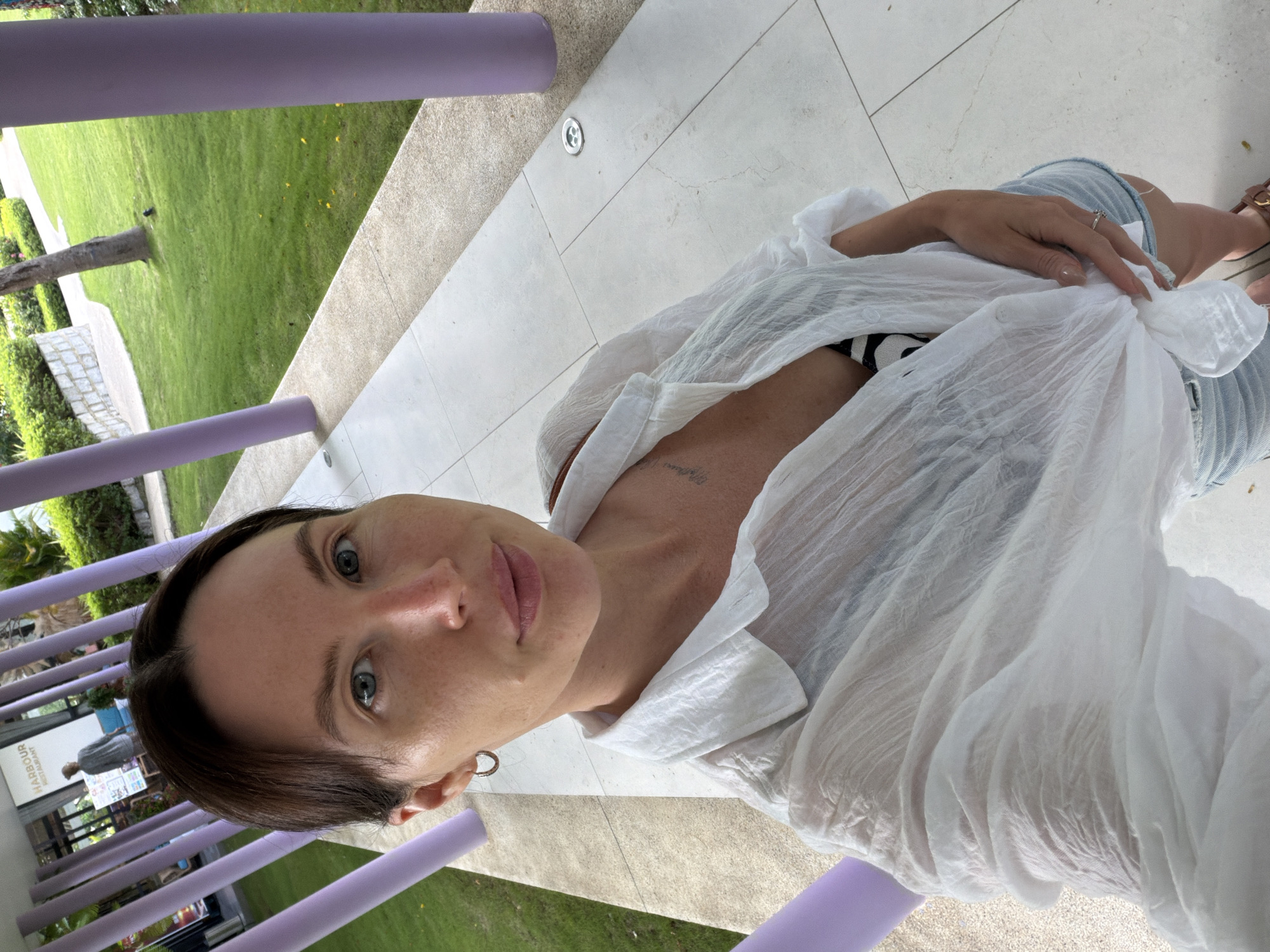 A woman is taking a selfie outdoors on a paved walkway with purple posts and a grassy area to the right. She has light skin, brown hair pulled back, and is wearing a white open-collared shirt tied at her waist, a black and white striped bikini top, and denim shorts. She has a tattoo on her chest and is wearing a hoop earring and a ring. In the background, there is a sign that reads "HARBOUR RESTAURANT". The location is Ninh Hoa Ward, Dong Hoi City, Vietnam.
