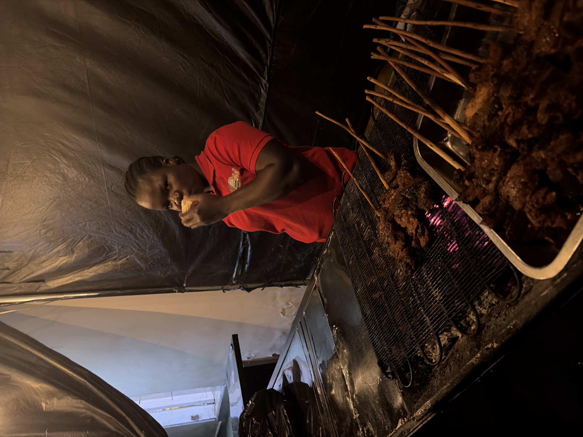 A young woman wearing a red polo shirt with a logo is eating a piece of food while standing in front of a grill in what appears to be an outdoor food stall. The grill is covered with meat skewers, some of which are actively cooking over glowing coals. The background is draped with dark fabric, creating a stark contrast with the bright light coming from an opening on the left side of the frame, revealing a partially visible white wall. The overall scene suggests a street food vendor preparing and serving food. The image is cropped to focus on the woman and the grilling area, with the city of Jos, Nigeria, being the presumed location.