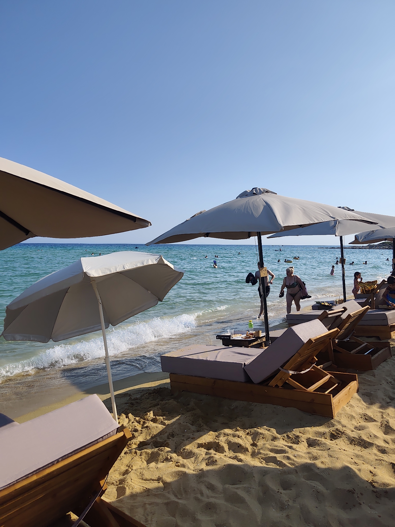 The image depicts a sunny day at a beach in Nea Peramos, Greece. The foreground is dominated by several empty, well-maintained lounge chairs and large umbrellas, indicating a beach club or resort setting. The chairs are made of wood with gray cushions. In the middle ground, the turquoise sea meets the sandy shore with gentle waves. Several people are visible in the water, some swimming and others enjoying the sea. A few individuals are walking along the waterline or entering the water. The background shows a clear blue sky. The overall impression is one of a relaxing summer day at a popular beach destination.