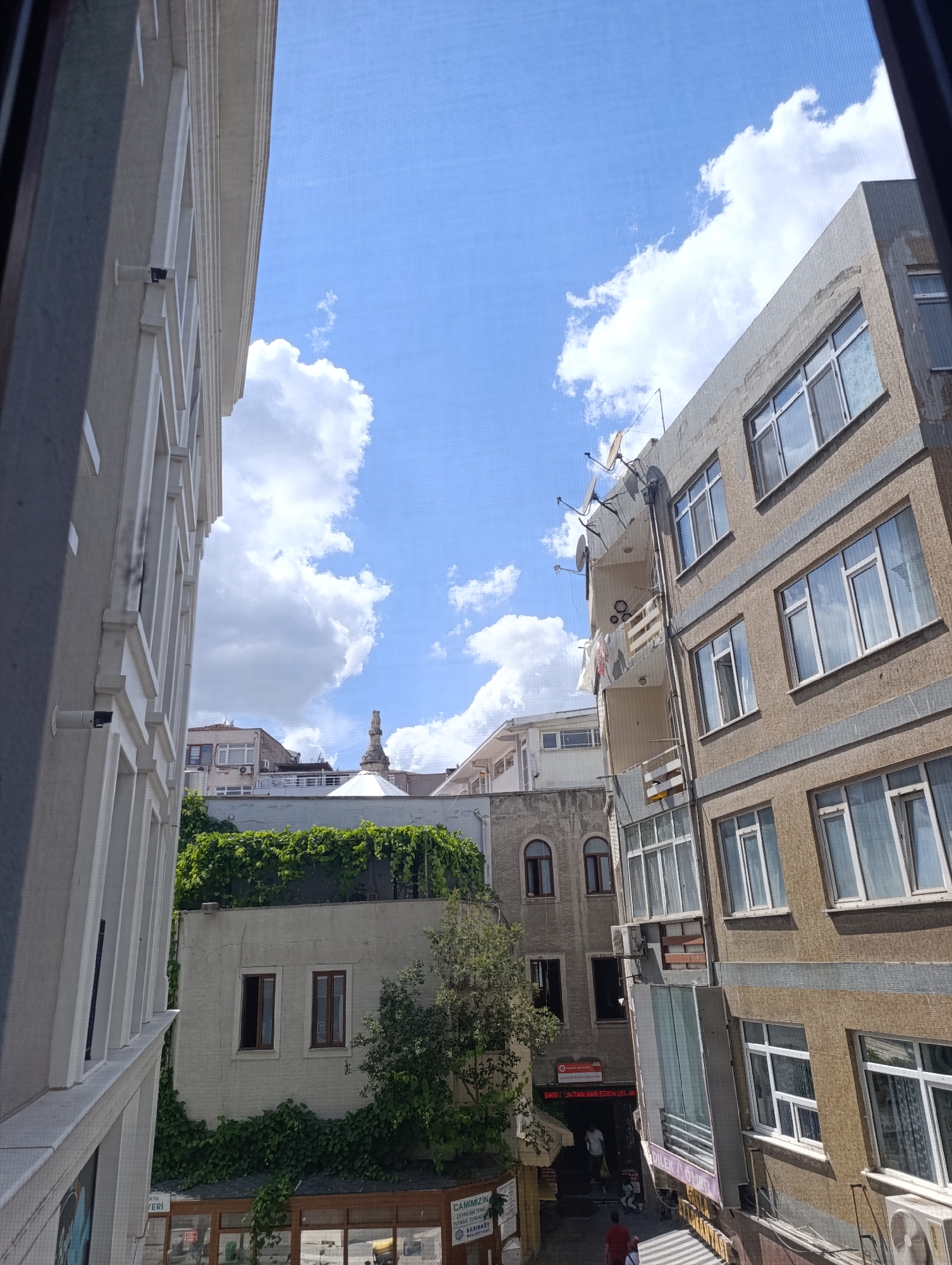 The image shows a view looking upwards and outwards from a building, capturing a section of the urban environment of Bakırköy, Turkey. On the left, a portion of a modern building with white decorative elements and windows is visible, extending vertically. To the right, a tan-colored apartment building with multiple floors and numerous windows is prominently featured. This building has a textured facade and several satellite dishes mounted on its exterior.

In the mid-ground, a smaller building is covered in lush green ivy, with two dark-framed windows visible on its upper level. Further back, a minaret-like structure, possibly part of a mosque, rises above the other buildings. The sky is a bright blue with scattered white cumulus clouds.

At the street level, the entrance to a building with a sign reading "CAMİMİZİN" is partially visible. There are a few people walking in the alleyway between the buildings. The overall scene depicts a dense urban setting with a mix of architectural styles.
