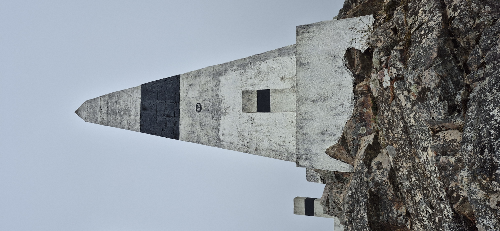 The image shows a concrete obelisk with black and white bands, situated on a rocky outcrop under an overcast sky. The obelisk is roughly triangular in shape and tapers to a point. There is a rectangular indentation with a black band on its face, and the number "1856" is inscribed in a circle to the left of this indentation. A smaller, similar concrete marker with black and white bands is visible to the left of the obelisk, also on the rocky terrain. The rocks are varied in color and texture, with patches of lichen and sparse vegetation. The scene appears to be a landmark or monument in an outdoor, elevated location. This monument is located at Monte da Madalena in Póvoa de Varzim, Portugal, and it marks the geographical location of the city.