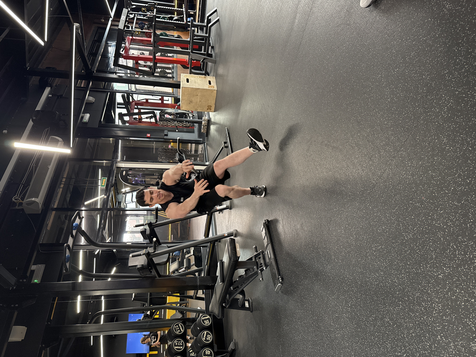 An indoor, eye-level shot captures a man in a modern gym, performing an exercise with his body angled, suggesting a dynamic movement or stretch. The image is rotated 90 degrees counter-clockwise. The man is wearing a black tank top, black shorts, and black and white athletic shoes. He has short, dark hair and a focused expression with a slight smile. His left leg is extended upwards, and his hands appear to be near his chest or waist.
The gym features dark walls and structural elements, accented by bright, linear fluorescent lighting strips on the ceiling and walls. Various pieces of gym equipment are visible, including weight racks, benches, and a wooden plyometric box labeled "20" and "30." A set of dumbbells is stacked in the foreground, with labels "ZIVA," "27.5," "50," and "48" clearly visible on different weights. The floor is a dark gray rubber material speckled with white. In the background, through glass partitions or reflections, more gym equipment and structures can be seen. A green "ВЫХОД" (EXIT in Russian) sign is visible high on a wall. A reflection of another person, possibly the photographer, is faintly visible in a screen or reflective surface near the dumbbells. The overall atmosphere is well-lit and active.