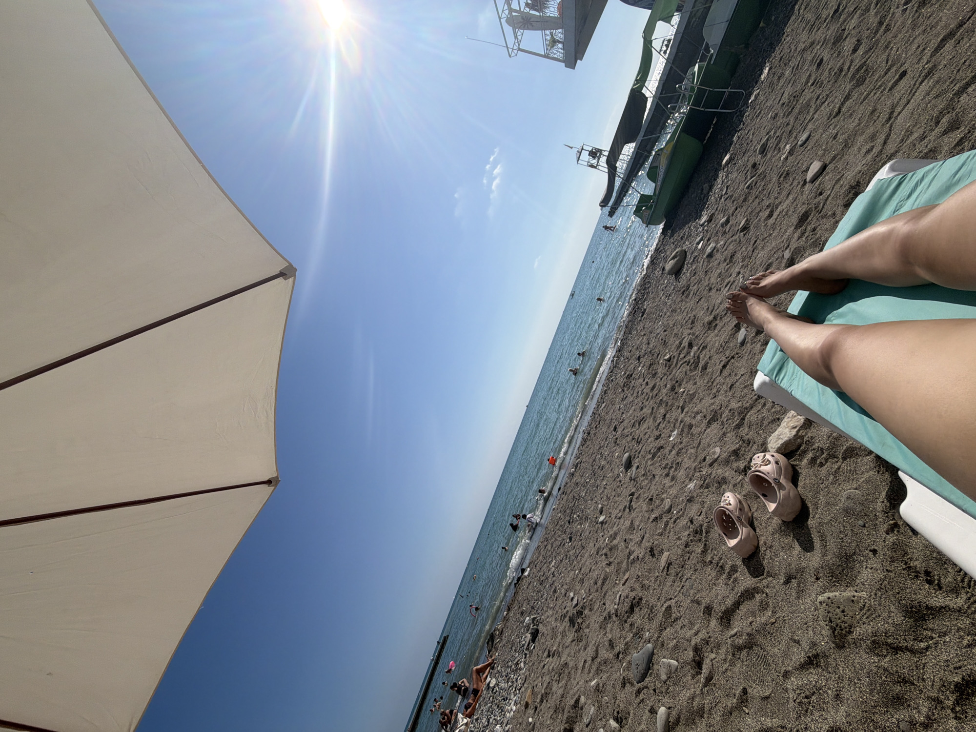 A low-angle, wide shot captures a sun-drenched beach scene with a clear blue sky and bright sunlight. In the foreground, a person's legs are stretched out on a turquoise lounge chair, with a white beach chair visible beneath. A pair of light pink clogs rests on the dark sand near the lounge chair. The beach is covered in dark sand and pebbles, sloping down to the calm, turquoise sea. Several people are visible in the water, swimming and wading. Along the shoreline, a few individuals are sunbathing on the pebble-strewn beach. In the background, two green motorboats with canopies are docked on the shore, with a lifeguard tower to the left of them. The sun's rays are prominently visible, creating a lens flare effect in the upper right corner of the image. This scene is consistent with a public beach in Sochi, Russia.