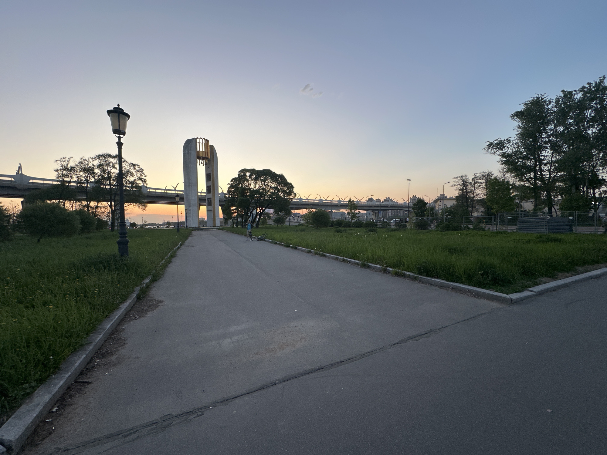 The image shows a park in Saint Petersburg, Russia at dusk. A paved path runs through the center, flanked by grassy areas with trees. A distinctive modern bell tower stands in the middle distance, with a highway overpass visible behind it. A street lamp stands along the path's edge. A person is visible in the middle distance, near the bell tower. The sky transitions from blue to orange near the horizon.