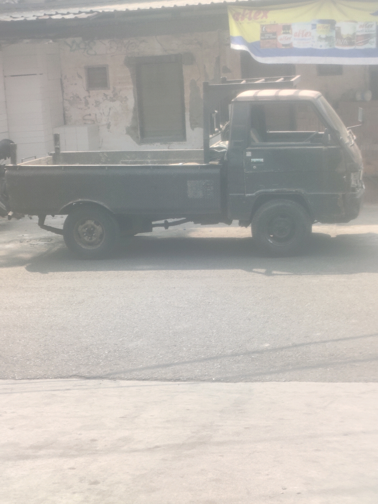 A dark-colored pickup truck is parked on an asphalt road in front of a building with a tiled roof. The building's exterior wall is weathered and shows signs of graffiti. A yellow banner with text is visible hanging above the truck. The truck has a single cab and an open bed, with its wheels visible. The scene appears to be in an outdoor urban environment.
