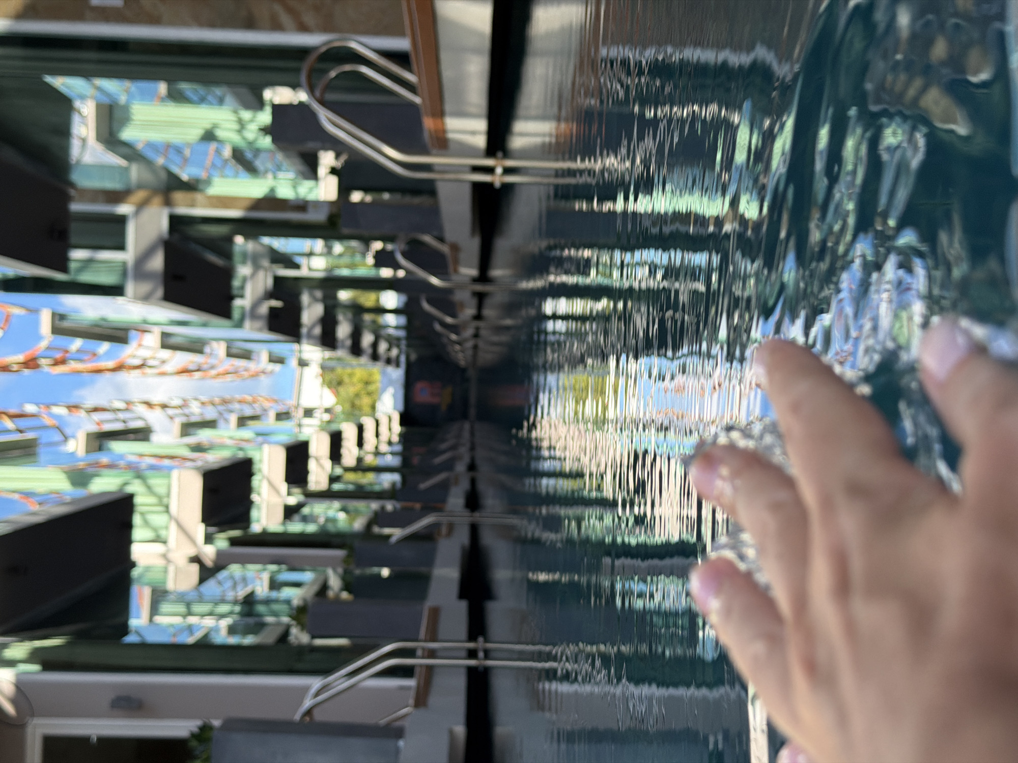 A person's hand, with manicured fingernails, reaches into the frame of the image, touching the surface of a rippling body of water. The water reflects distorted images of buildings and a blue sky. The buildings appear modern with large glass windows and a green hue. There are metal railings visible, suggesting this might be a swimming pool or a decorative water feature. The scene is outdoors, with natural light indicating daytime. The overall impression is one of a person interacting with water in an urban or resort-like environment. The image is oriented vertically, with the water filling the right side and the buildings the left.