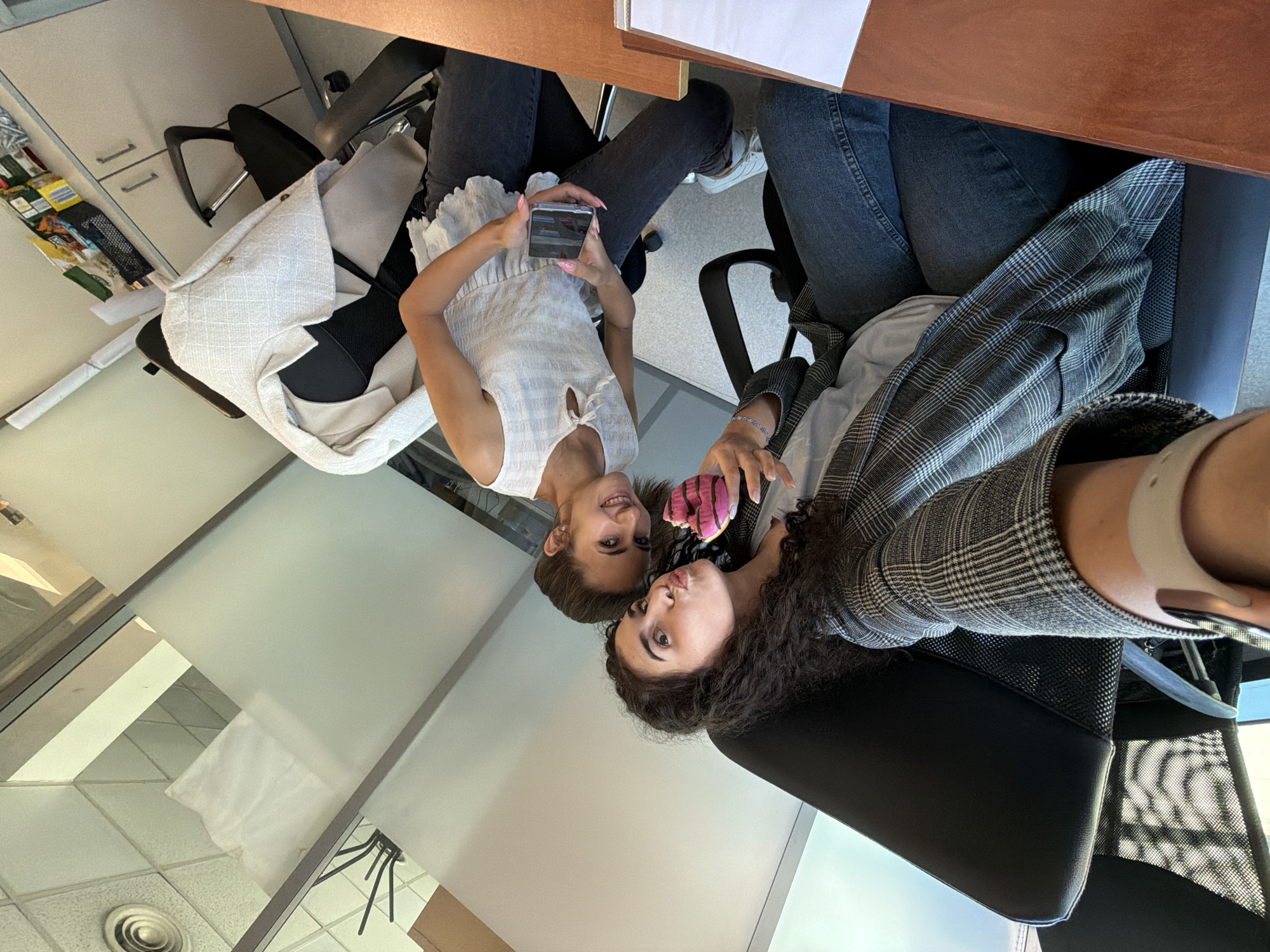 Two women are seated in office chairs, taking a selfie in an office environment. The woman on the left is closer to the camera and has dark curly hair. She is wearing a grey and white checkered blazer over a light-colored shirt and dark jeans. She is puckering her lips and holding a partially eaten pink frosted donut. The woman on the right has brown hair pulled back and is wearing a white ruffled top and dark jeans. She is holding a smartphone in her right hand, seemingly taking the picture. The background shows office furniture, including desks, chairs, and cabinets, with frosted glass partitions. The scene appears to be a typical office setting in Sofia, Bulgaria.