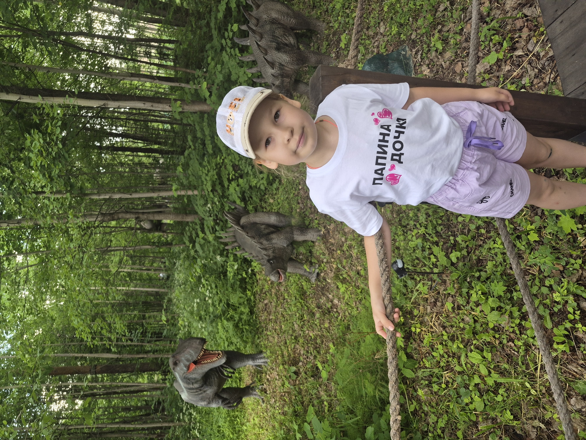 A young girl, approximately 6-7 years old, is standing in a wooded area, holding onto a thick rope. She is wearing a white t-shirt with the Russian text "ПАПИНА ДОЧКА" (Daddy's Daughter) printed in black, accompanied by pink heart graphics. She also wears light purple shorts and a white baseball cap with animal graphics. Behind her, several dinosaur replicas are placed among the trees and foliage. To her left, a T-Rex replica stands with its mouth open. To her right and slightly behind, two other dinosaur replicas, possibly Ankylosaurs, are visible. The environment appears to be a park or an outdoor exhibition, with a wooden pathway and a natural forest setting with deciduous trees and green undergrowth.