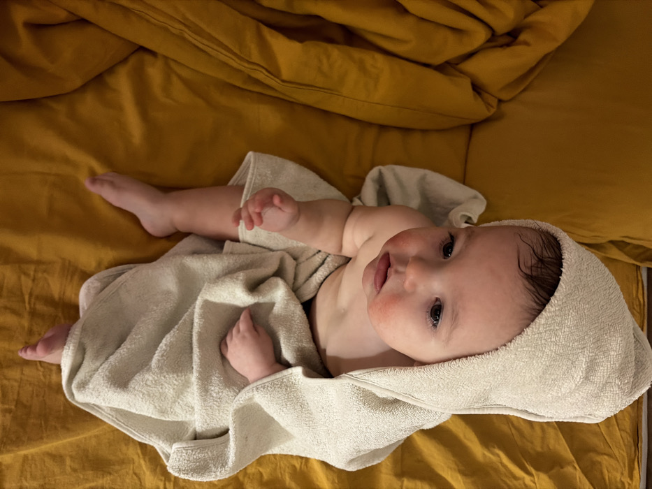A baby lies on a yellow bedsheet, wrapped in a light green hooded towel. The baby is positioned at an angle, looking upwards. Their tiny hands and feet are visible outside the towel. The skin on their cheeks appears slightly reddened. The bedsheet is slightly wrinkled. The lighting in the image is warm and soft.