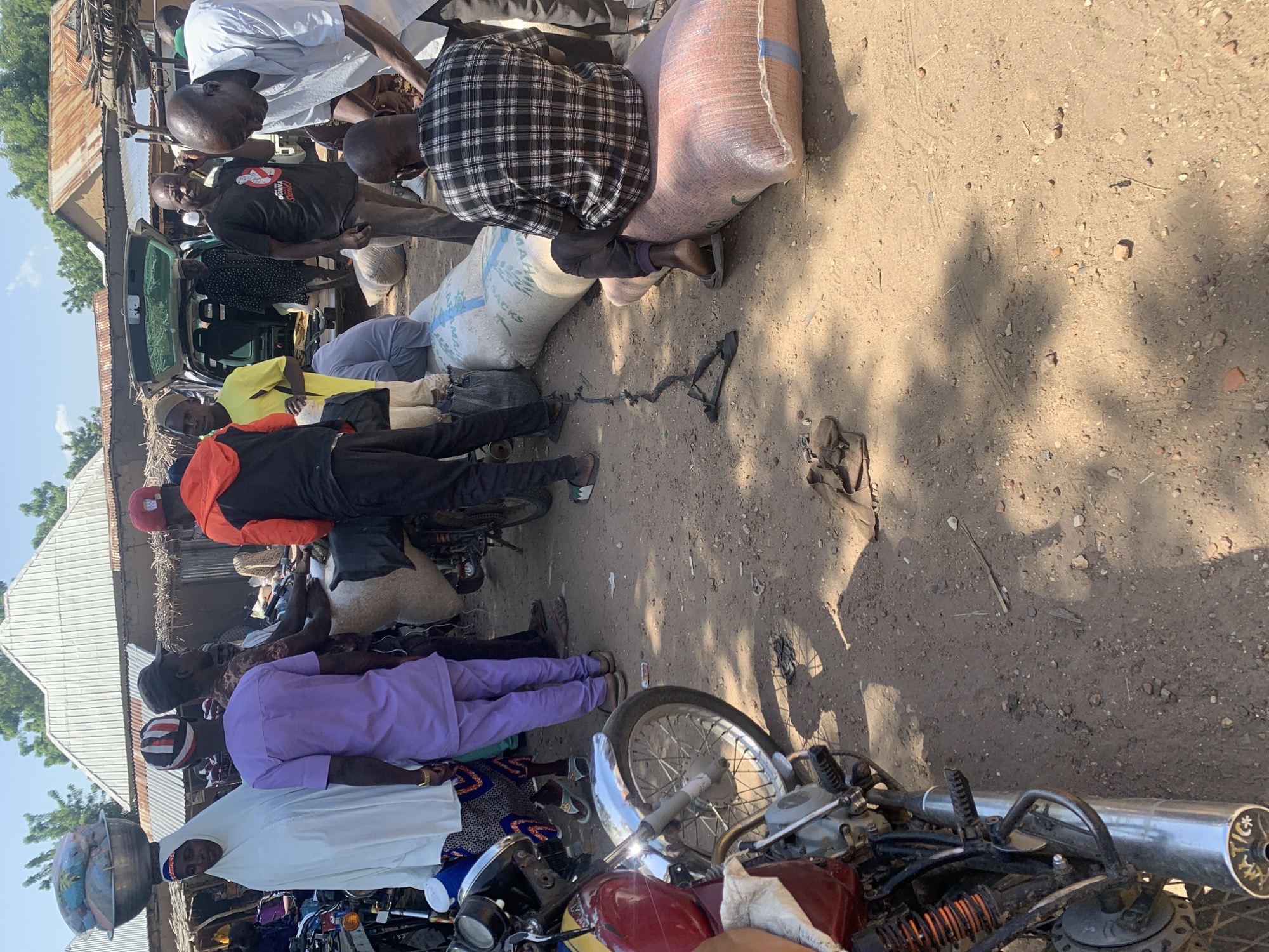 This media file depicts a scene in Boi A, Nigeria, with several individuals engaged in the loading or unloading of large sacks, likely containing goods, onto or from a vehicle. The overall setting appears to be an outdoor market or roadside area, characterized by a dirt ground, some rudimentary structures with corrugated metal roofs, and vegetation in the background.
The subjects are a group of men, some of whom are actively handling the sacks. One man wears a plaid shirt and is partially obscured by a large sack. Another man in a black t-shirt with a graphic on it appears to be directing or observing the activity. Several other men are visible in various states of participation, including one wearing a bright yellow shirt and another in a purple tunic. The presence of motorcycles in the foreground and background suggests transportation is a key element of the scene, likely used to bring the goods to this location or to transport them away.
The time of day appears to be daytime, as indicated by the strong shadows cast on the ground, suggesting bright sunlight. The weather seems to be warm and dry, consistent with an outdoor market environment in Nigeria. The mood seems to be one of industrious activity, with people focused on their tasks.
One of the sacks has some visible text on it, including what appears to be "SACKS" in large letters. Other markings on the sacks are less legible. The overall impression is of a bustling, working environment where goods are being moved, possibly for trade or distribution.
