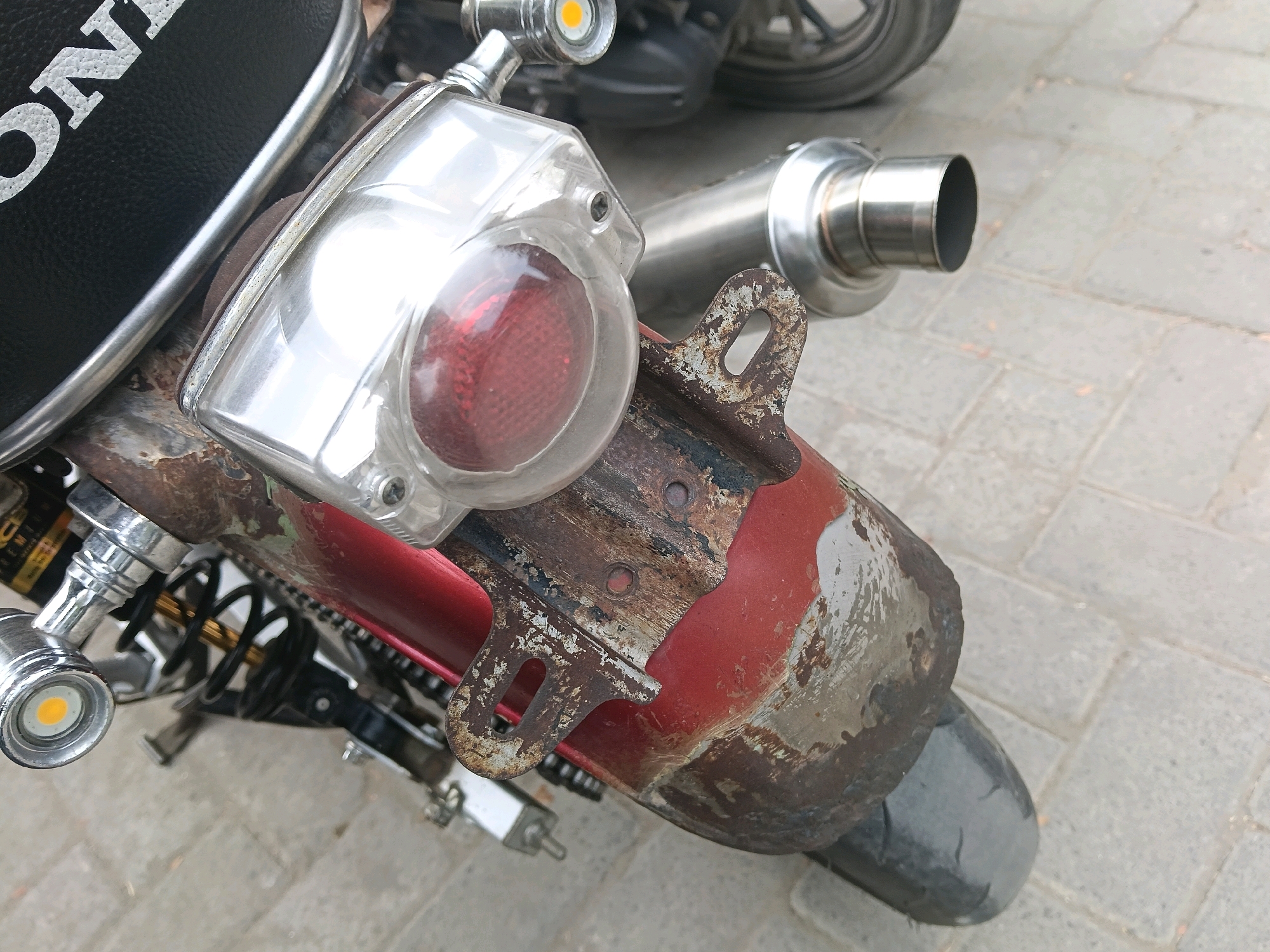 The image shows a close-up view of the rear fender and tail light of a vintage motorcycle, with the word "HONDA" visible on the seat. The motorcycle appears to be in a state of disrepair, with rust and peeling paint on the fender. A chrome exhaust pipe is also visible. The background consists of a paved surface, likely a sidewalk or street, with a gray, rectangular brick pattern. The lighting suggests it is daytime. The image was taken in Balun, Indonesia, in front of a Honda motorcycle parked on a paved walkway.