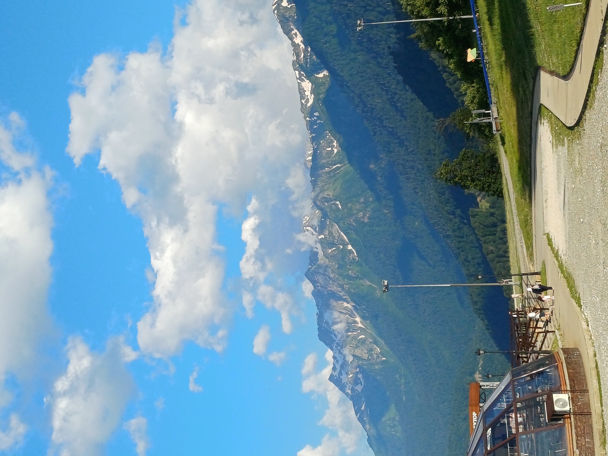 The image depicts a bright, daytime scene with a focus on a mountain range and a landscaped area in the foreground. The sky is a vibrant blue with scattered, fluffy white clouds. The mountains in the background are majestic, with their peaks dusted with snow, contrasting with the lush green slopes covered in dense forest. The lower slopes are a dark, deep green, indicating a thick canopy of trees.
In the mid-ground, a paved pathway curves through a grassy area. There are several lampposts scattered along the pathway, with one prominent tall lamppost with multiple lights standing out in the right half of the frame. A few trees are visible, with one distinct tall evergreen tree towards the right. A blue barrier with some signage is also present.
The foreground features a modern, glass-enclosed structure with a brick base, possibly a cafe or visitor center. The structure is partially visible on the left side of the image. Next to it, a wooden fence with a railing follows the curve of the pathway. Several people are visible walking along the pathway and near the fence. Two individuals, a woman in a hijab and a child in pink, are walking together on the paved path. Other figures appear to be in groups or walking individually, suggesting a public space with recreational activity. The ground in the foreground is covered with gravel and paved sections. The overall impression is a scenic location in a mountainous region, likely a resort or park area, potentially in Estosadok, Russia, given the context.