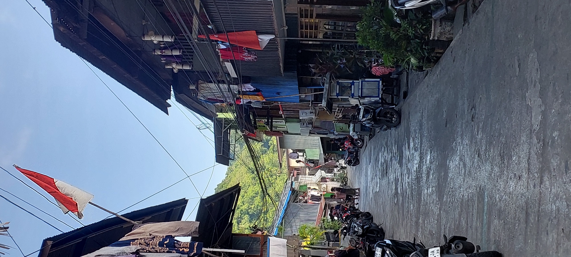 The image depicts a narrow street lined with multi-story dwellings and businesses, likely in a densely populated urban or semi-urban area. The architecture appears utilitarian, with corrugated metal roofs and various building materials evident. Laundry is hung from clotheslines strung between buildings, suggesting residential use. Several motorcycles are parked along the street, indicating a common mode of transportation. In the background, a lush, green hillside is visible, suggesting proximity to nature. The sky is clear and blue. The scene is bathed in natural light, casting shadows on the ground. The street surface is paved and appears somewhat damp or reflective in places. A flag, likely the Indonesian flag (red and white), is prominently displayed from one of the buildings, suggesting a patriotic or celebratory context. People are present on the street and on balconies, engaged in various activities, though specific interactions are not clearly discernible. The overall impression is one of a vibrant, lived-in community.
