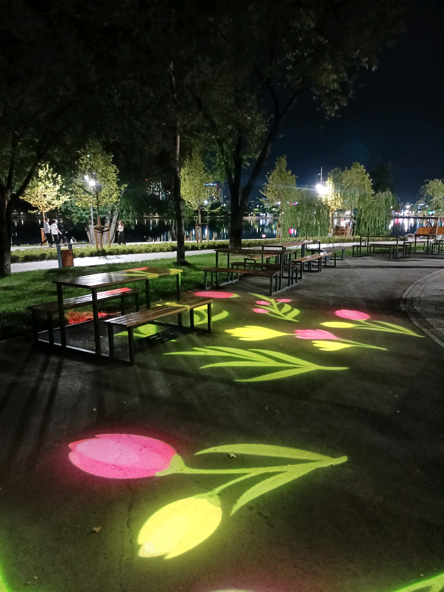 In a nighttime scene in an urban park, picnic tables and benches are illuminated by projections of colorful tulips on the dark asphalt path. The park features a body of water in the background, with trees and lights lining its edge. Several trees with illuminated foliage stand on a grassy area to the left of the path. Three people are walking along the paved pathway in the background, and another person is standing near a bench.