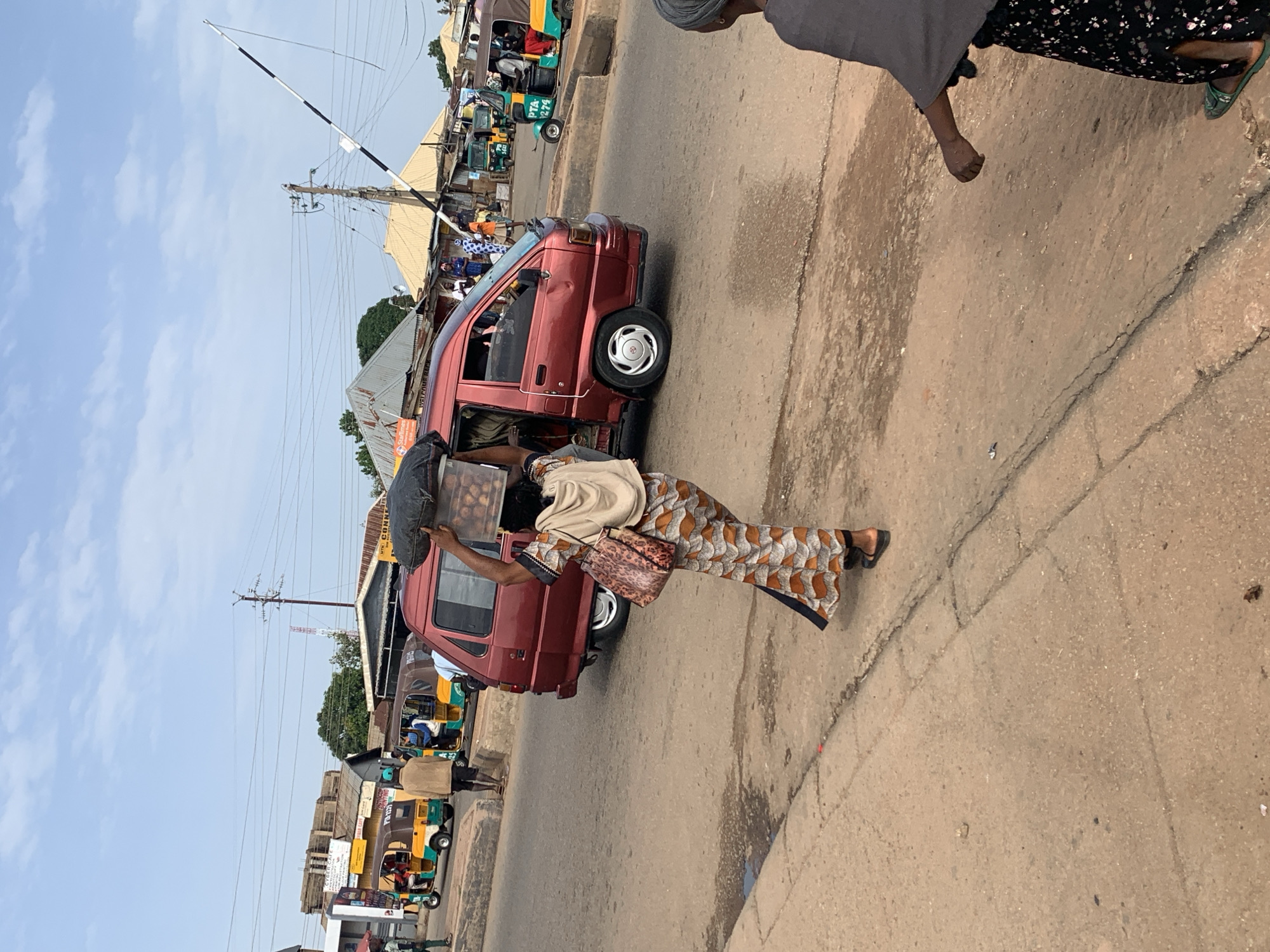 A woman in a patterned dress and headscarf carries a clear plastic container filled with goods on her head. She walks along a paved road in Gura Top, Nigeria, on a cloudy day. Behind her, a red van is parked, and several auto-rickshaws, some with passengers, are visible on the road. Buildings and utility poles line the street. Another woman, wearing a grey shawl and floral skirt, is seen from the side in the foreground, walking away from the camera.