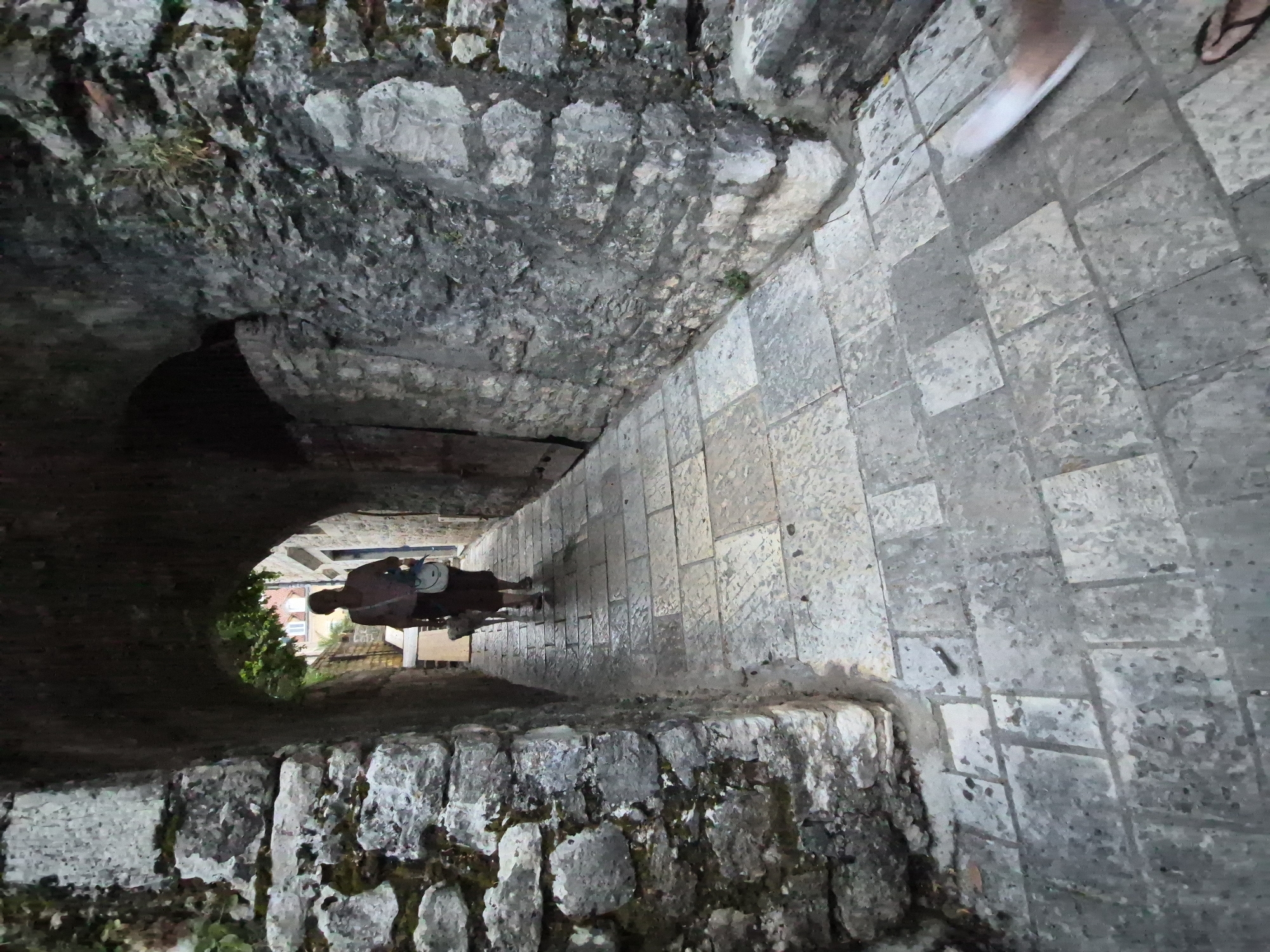 The image depicts a narrow, stone-paved pathway winding through an ancient stone structure, likely a fortress or old town in Herceg Novi, Montenegro. The pathway is lined by rough-hewn stone walls, some of which are partially covered in moss and vegetation, suggesting an older, possibly damp environment. On the right side of the pathway, a person's feet are visible, one wearing a sandal, indicating the current time of day is likely daytime. The lighting suggests it might be late afternoon or early evening, with a slightly diffused light. The pathway appears to be leading into a darker, arched opening in the stone structure on the left, offering a glimpse of foliage and buildings beyond. In the center of the pathway, a dark, possibly metallic object stands upright, resembling a cannon, further hinting at the historical and defensive nature of the location. The overall atmosphere is one of historical depth and weathered resilience, with the stone surfaces showing signs of age and exposure to the elements.