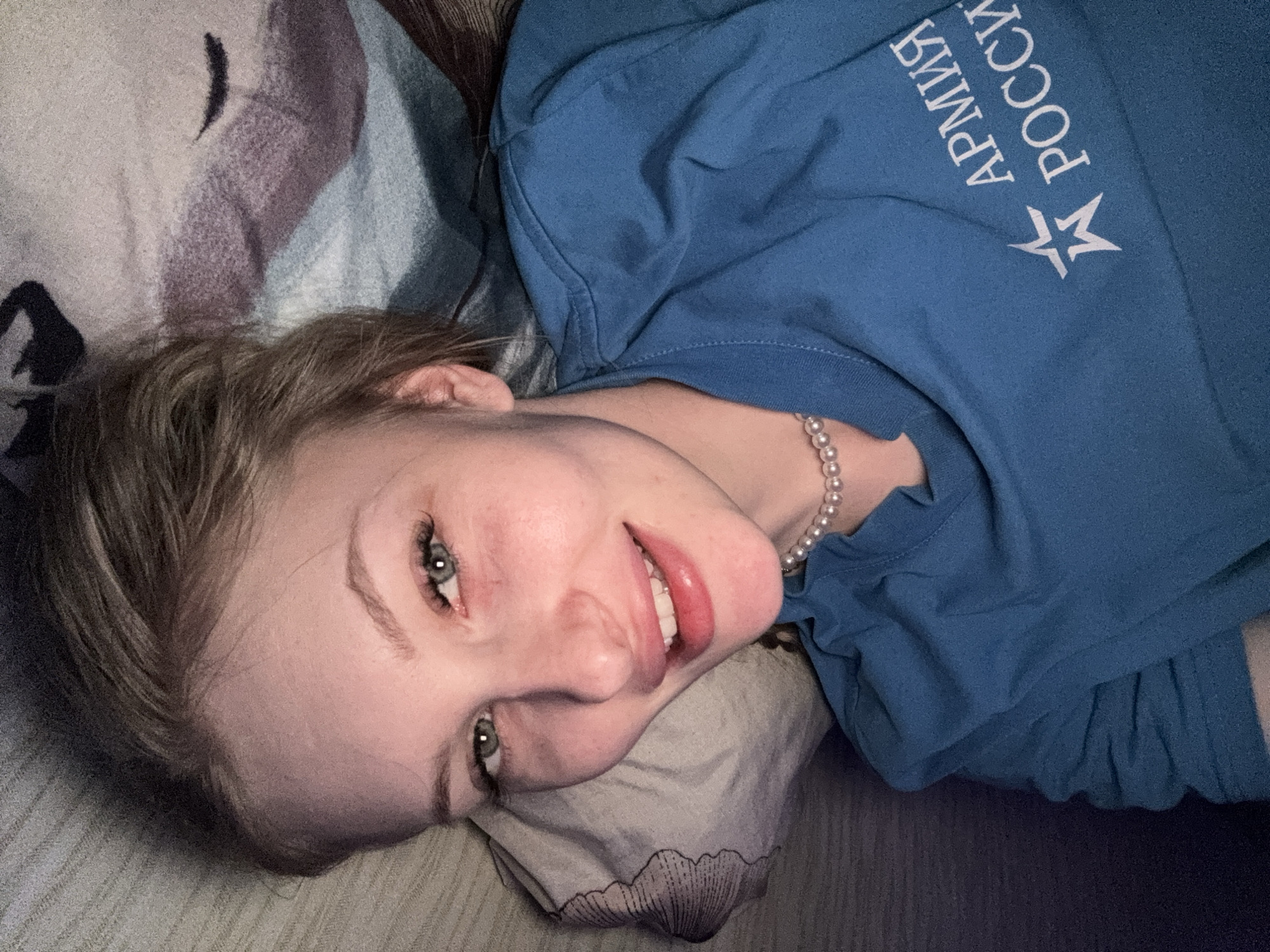 A young woman is captured in a close-up, indoor shot, lying down and looking directly at the camera with a cheerful, relaxed smile. Her head rests on a pillow with a subtle patterned design. She has light hair, visible around her face, and striking blue-green eyes. Her lips are slightly parted, showing a glimpse of her teeth, and appear to have a glossy finish.
She is wearing a string of white pearl beads as a necklace and a blue short-sleeved t-shirt. On the front of the t-shirt, mirrored white text and a star symbol are visible. When viewed correctly, the text reads "АРМИЯ РОССИИ," which translates to "Army of Russia." The star emblem, a common military symbol, accompanies the text. The background consists of textured fabrics, including a patterned bedsheet or blanket and a darker, plain material. The lighting suggests an indoor setting with soft illumination. The overall mood conveyed is one of contentment and approachability. The image context indicates the location is Kondratovo, Russia.