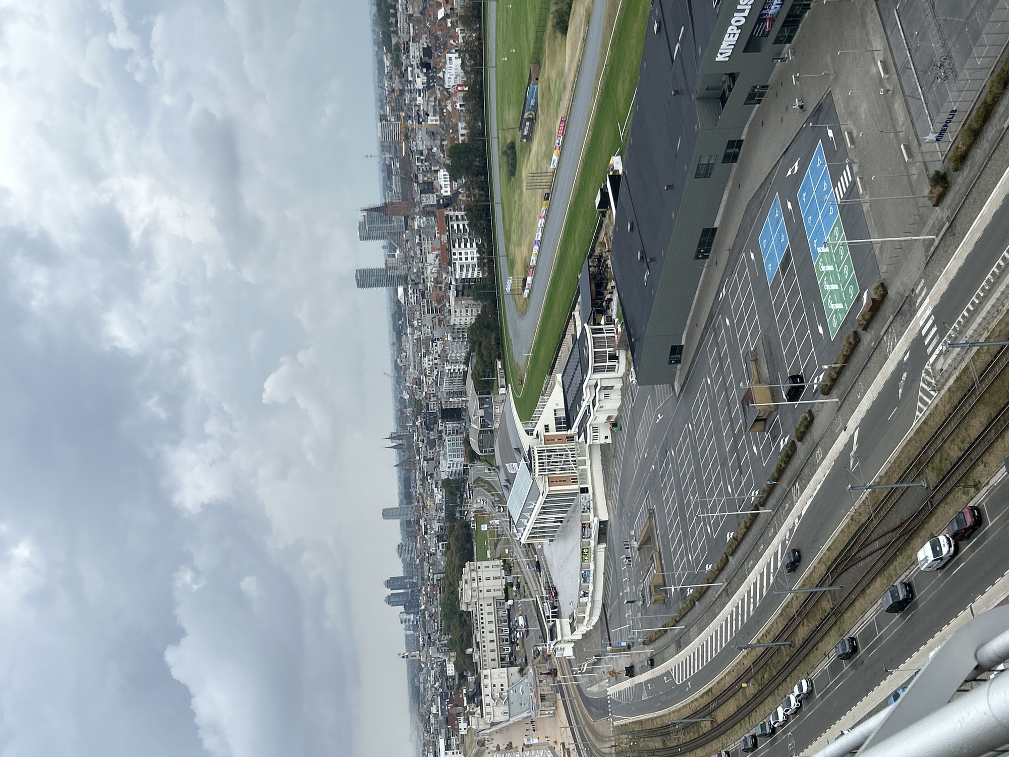An aerial, high-angle, full shot captures a cloudy day in Ostend, Belgium, looking inland from the coast. In the foreground, a modern, dark-gray building with "KINEPOLIS" signage is prominent, featuring a large parking lot with designated disabled parking spaces marked in blue and green. Adjacent to the parking lot is a paved area with benches and a few bicycles parked, also bearing "KINEPOLIS" branding. To the left, tram tracks run alongside a road with several cars and vans parked or driving. Further back, a white building with an arched roof and a facade adorned with red bricks sits beside a grassy area that transitions into a horse racing track. In the middle ground, a collection of mid-rise and high-rise buildings forms the cityscape, with a mix of residential and commercial structures, some with distinctive architecture. The background is dominated by a vast expanse of overcast sky, with large, textured clouds filling the frame. The overall impression is an urban environment with a blend of modern entertainment facilities, residential areas, and infrastructure, set against a typically cloudy coastal sky.