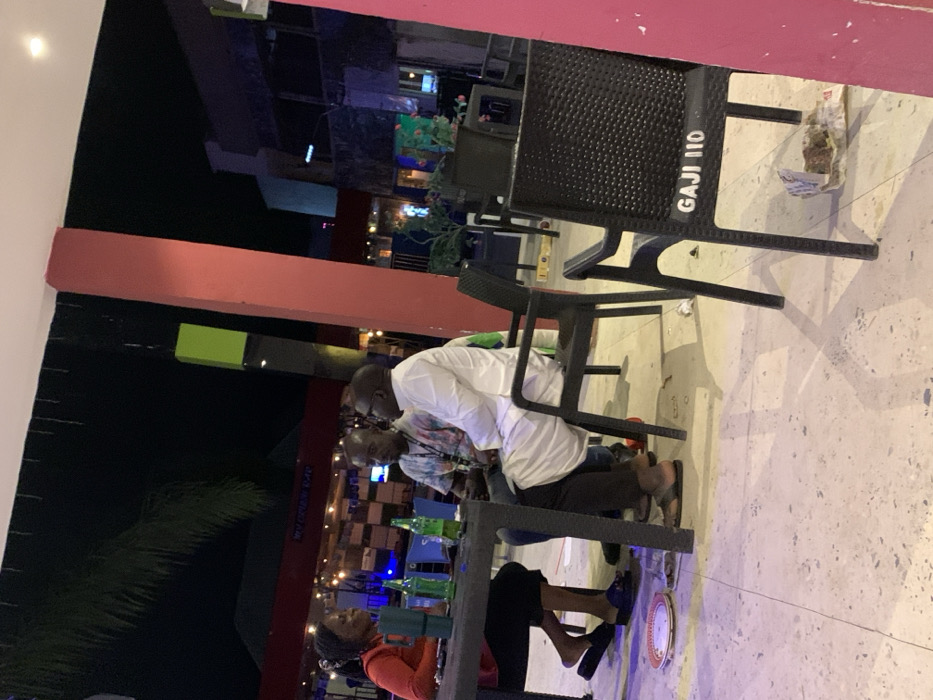 The image depicts an outdoor seating area at night in a commercial establishment in Akwanga, Nigeria. Three individuals are seated around a dark rectangular table on a light-colored tiled patio.
A female, wearing a red long-sleeved top, dark lower garment, and dark slippers, is positioned on the left, facing right. Her hair is styled in dreadlocks or braids. To her right, a male wearing a multi-colored tie-dye shirt, dark pants, and light brown sandals sits, facing right. He has short dark hair and wears glasses. To his right, a second male, dressed in a white traditional long garment (possibly an agbada or kaftan) over dark pants, and dark footwear, sits facing left, towards the other two. He also has short dark hair and wears glasses.
On the table are several green-bottled beverages and a dark insulated mug. All three individuals occupy black plastic chairs. An unoccupied black plastic chair in the foreground displays "GAJI 110" in white lettering on its backrest. On the tiled floor beneath the table, a white plate with a red rim is visible. Near the unoccupied chair, discarded materials, including a wrapper and food scraps, are present.
The background is dark, indicating a nighttime setting, illuminated by artificial lights. A prominent red structural pillar is visible to the right and center. A green rectangular light fixture or decoration is seen above the central pillar. Further in the background, outlines of buildings with various lights are discernible, including a partially visible sign that reads "WONDER BAR." Palm fronds are silhouetted against the dark sky at the top left.