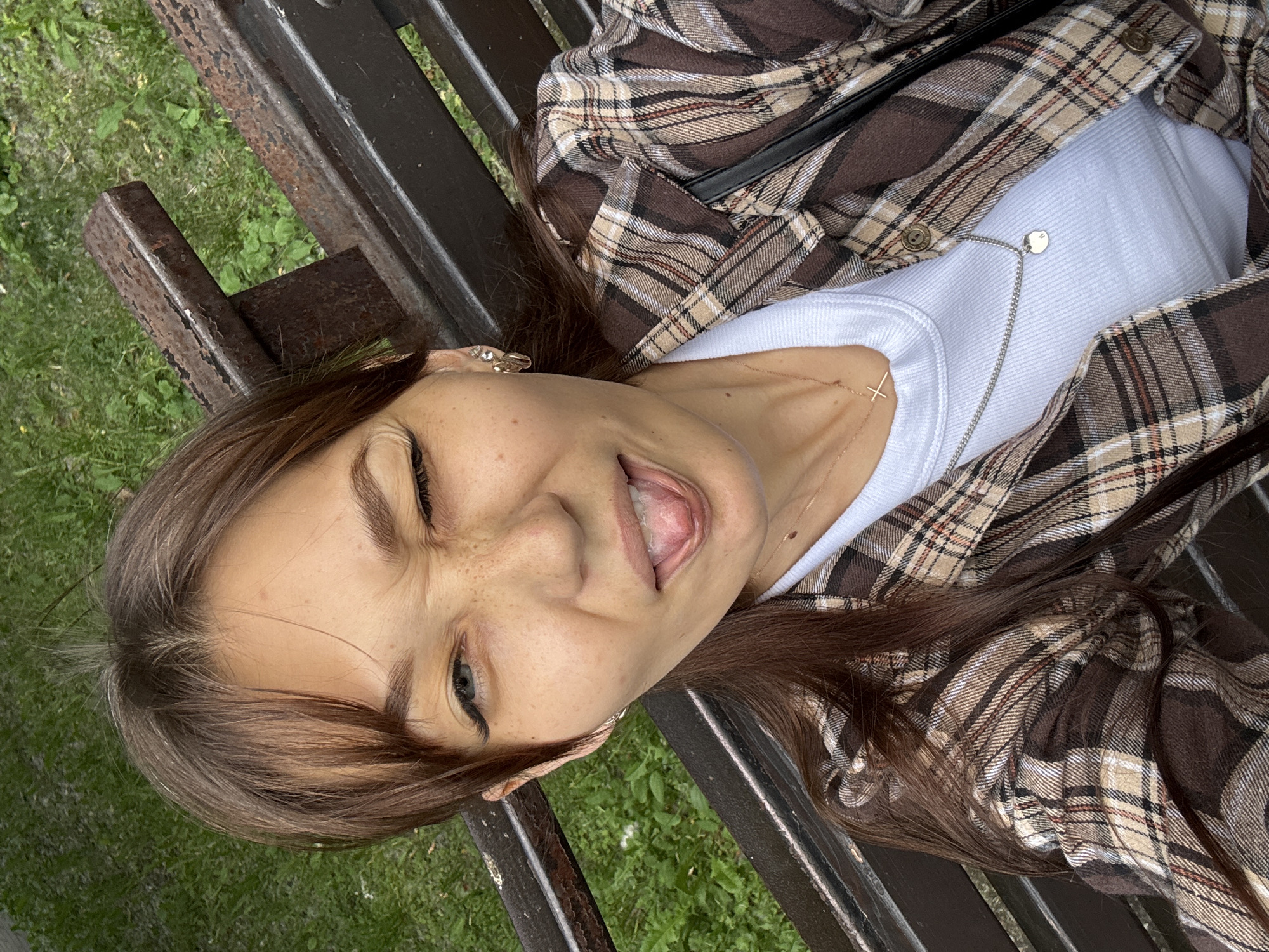 This is a vertical, eye-level selfie of a young woman with long brown hair, lying down on a park bench. She is wearing a brown and beige plaid shirt over a white tank top and a delicate silver necklace with a small heart pendant. Her eyes are closed, and her mouth is open with her tongue visible, giving a playful or excited expression. She has light skin with freckles on her cheeks and nose. The bench she is on appears to be made of dark wood or metal, with some visible rust. Behind the bench, out of focus, is lush green grass and some foliage, suggesting an outdoor setting on a bright day. The overall impression is of a spontaneous and joyful moment captured in a natural environment. The location context indicates this photo was taken in Novokuznetsk, Russia.