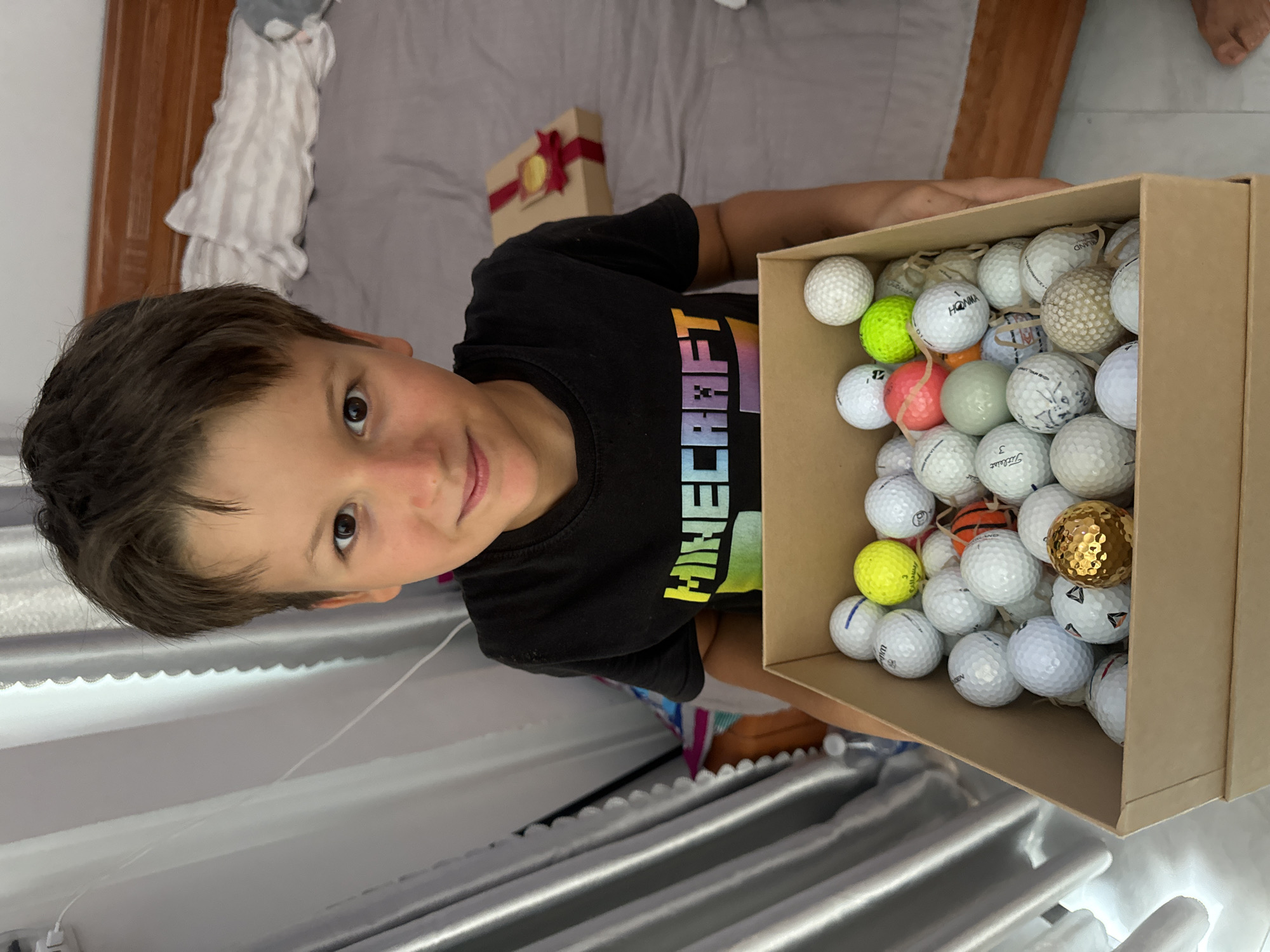 A young boy in a black Minecraft t-shirt is holding a box full of golf balls. The box contains various golf balls, some with brand names and markings, including a gold-colored ball. In the background, there is a bed with a small, wrapped gift box with a red ribbon. The location appears to be indoors, possibly a home, in Mũi Né, Vietnam.