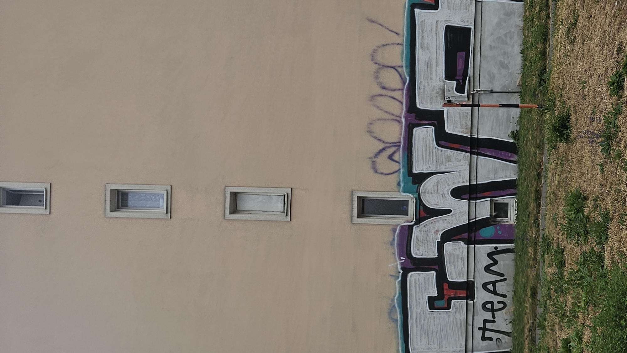 The image shows a building with a light brown facade, featuring three narrow, vertically oriented windows stacked one above the other on the upper section of the wall. Below these windows, a large graffiti piece covers the lower part of the wall. The graffiti consists of stylized letters in white with black outlines, and some sections filled with purple and teal colors. To the left of the main graffiti, the word "TEAM" is written in black block letters. Above the main graffiti, faint purple graffiti marks are visible. A small, dark window is situated within the lower graffiti section, and another small window is present to the right of the "TEAM" text. In front of the wall is a patch of dry grass and some sparse green vegetation. A thin, orange and black striped pole is also visible in front of the wall, positioned to the right of the graffiti. The scene appears to be in an urban environment. Based on the architectural style and the graffiti, this image is likely from a residential area in Prešov, Slovakia, a city known for its urban art scene.