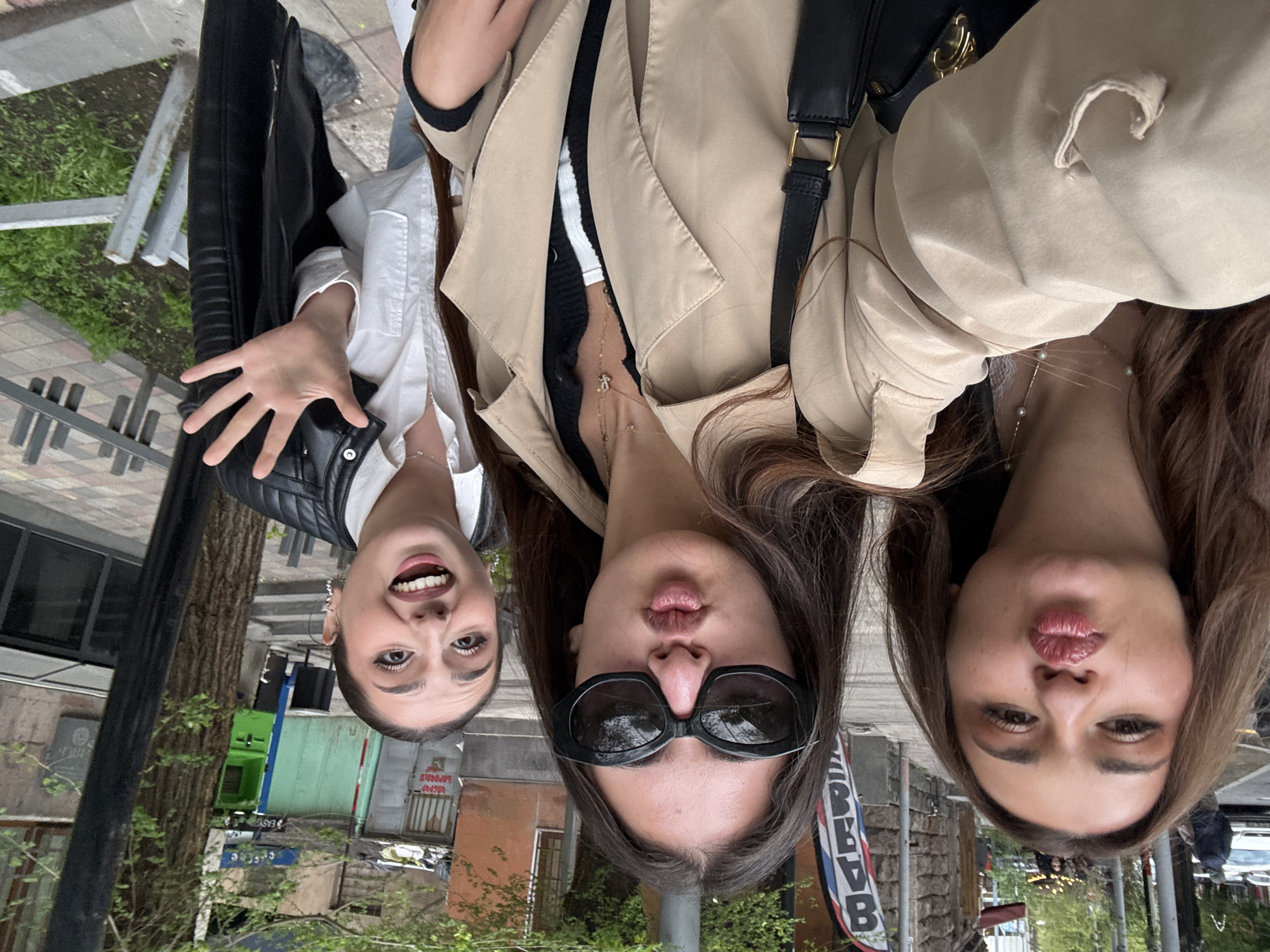 Three young women are depicted in a playful outdoor selfie, with the image rotated 90 degrees clockwise. They are looking upwards towards the camera with expressive faces.
The woman on the left has dark hair, pulled back, and sports a wide, open-mouthed smile, revealing her teeth. She wears a white collared shirt beneath a black leather jacket, and her left hand is raised towards the camera, fingers spread. The central woman has dark hair and dons dark sunglasses, puckering her lips into a 'kissy face.' She is dressed in a beige trench coat over a black and white striped top, accessorized with several delicate gold necklaces, one featuring a small, cross-like charm. The woman on the right, with brown hair, also makes a 'kissy face' with puckered lips. She wears a beige trench coat and a gold layered necklace adorned with small pearls.
The setting appears to be an urban street or sidewalk during daytime, characterized by soft, even lighting. The background includes concrete pavement, green foliage from trees and bushes, various buildings (some with brick facades, others with glass windows), a street pole, and metal barriers.
Visible text includes "EASY" and an arrow on a green sign located behind the leftmost woman. A vertical banner positioned behind the middle and right women displays Cyrillic characters, which appear to spell "PЯВАВ" or a similar word, suggesting a local business or advertisement. The overall impression is one of casual camaraderie and cheerful posing.