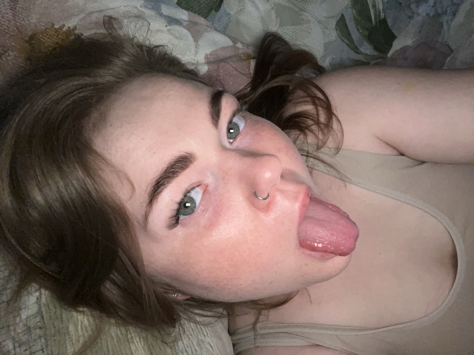 A close-up, angled shot of a woman lying on a patterned surface, possibly a bed or couch. She has light skin, brown hair, and green eyes. She has a nose ring and is sticking her tongue out. She is wearing a light-colored tank top. The background is blurred. There is no evidence in the image to suggest this was taken in Chaikovsky, Russia, or any other location.