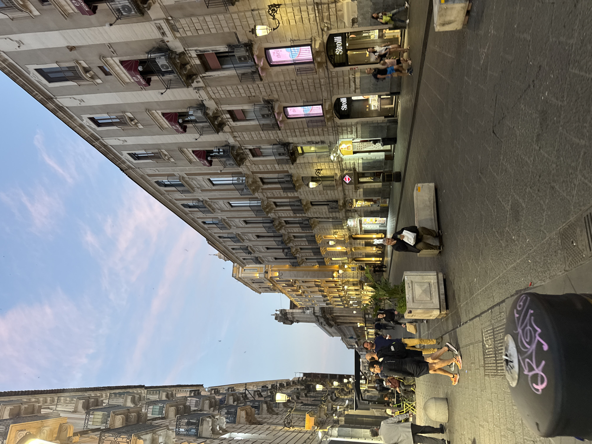 The image depicts a street scene, likely taken at dusk or in the early evening, with buildings lining both sides of the street. The structures are multi-storied with architectural details like windows with small balconies and awnings. Some shops have illuminated signs. There are people walking along the street, some standing, some sitting. The street is paved with stone. A streetlamp or bollard with graffiti is prominently featured in the foreground. The sky is a mix of blue and pale pink, suggesting the time of day.