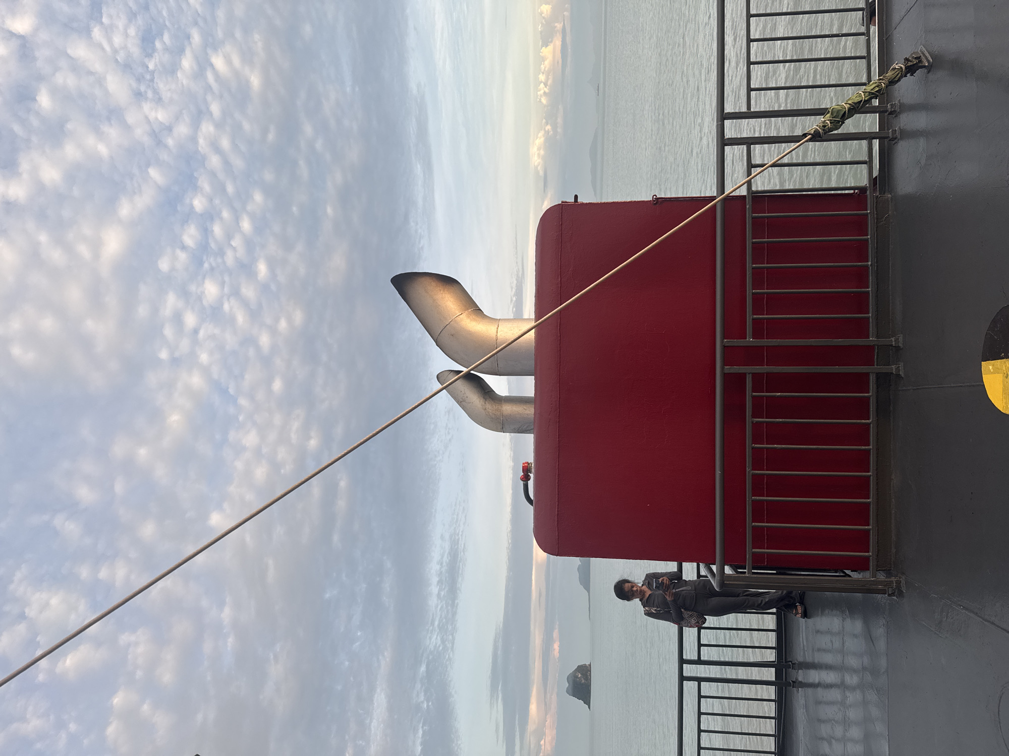 The image captures a view from a ferry deck with a woman standing near the railing and looking at her phone. In the background, the calm sea stretches to the horizon, with distant islands visible under a sky filled with scattered clouds. The ferry features a prominent red structure with two large exhaust pipes angled upwards. A thin rope extends diagonally across the frame, secured to the deck with a green-wrapped bracket. The woman is wearing dark, loose-fitting clothing. The overall scene suggests a journey at sea near Don Sak, Thailand, likely during dawn or dusk due to the soft lighting.